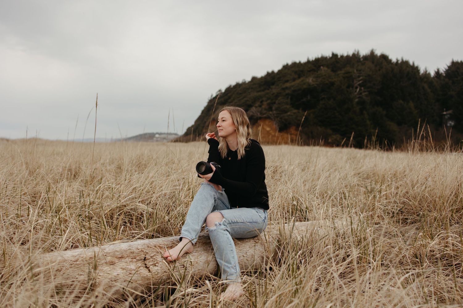A young woman sits on a log in a golden field, holding a camera on an overcast day near a forested hill.