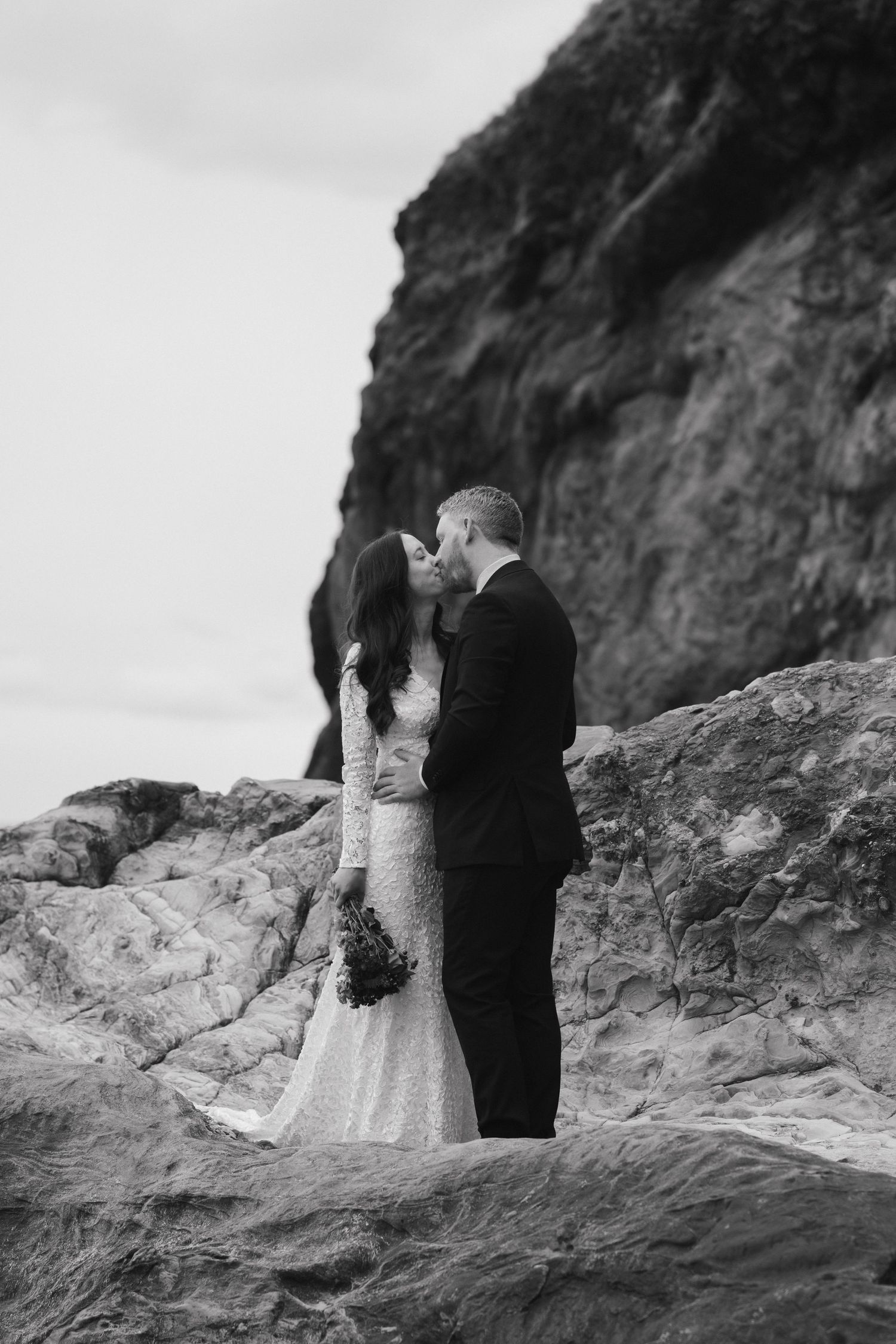 A romantic black and white photo of newlyweds embracing on coastal rocks.