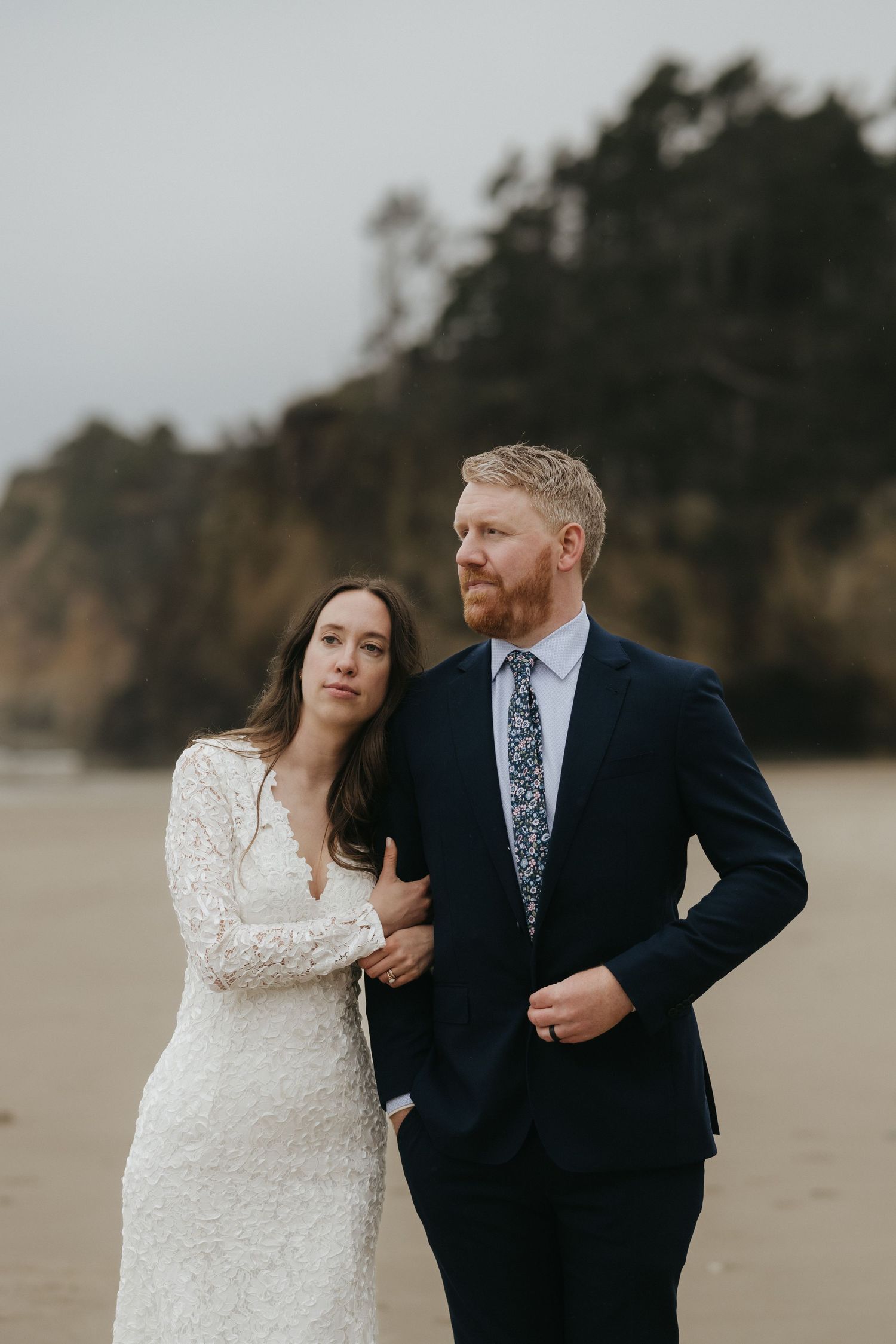 A couple in wedding attire embraces on a beach with dramatic cliffs in the background.