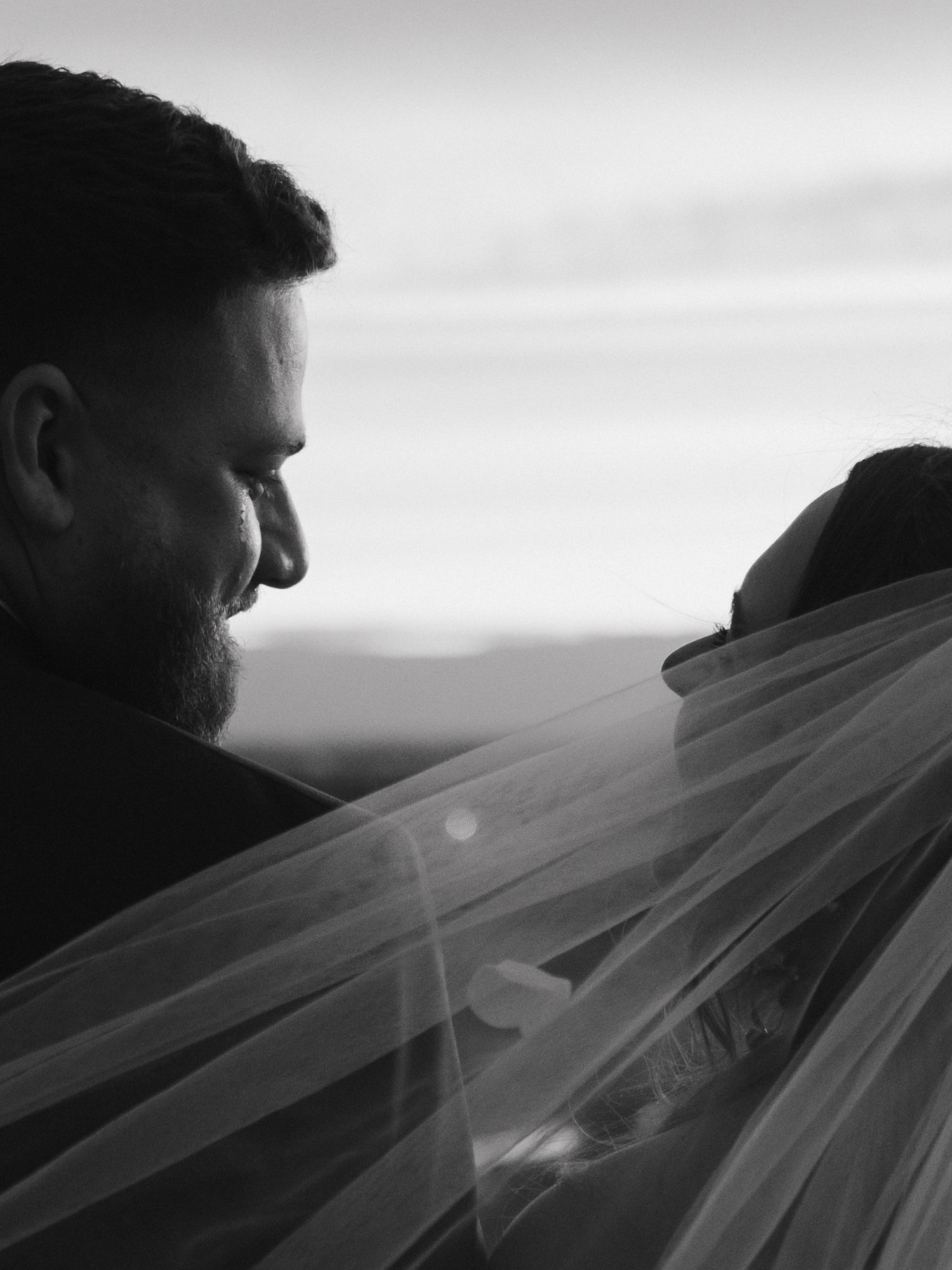 Artistic black and white close-up of flowing wedding veil against a sunset sky.