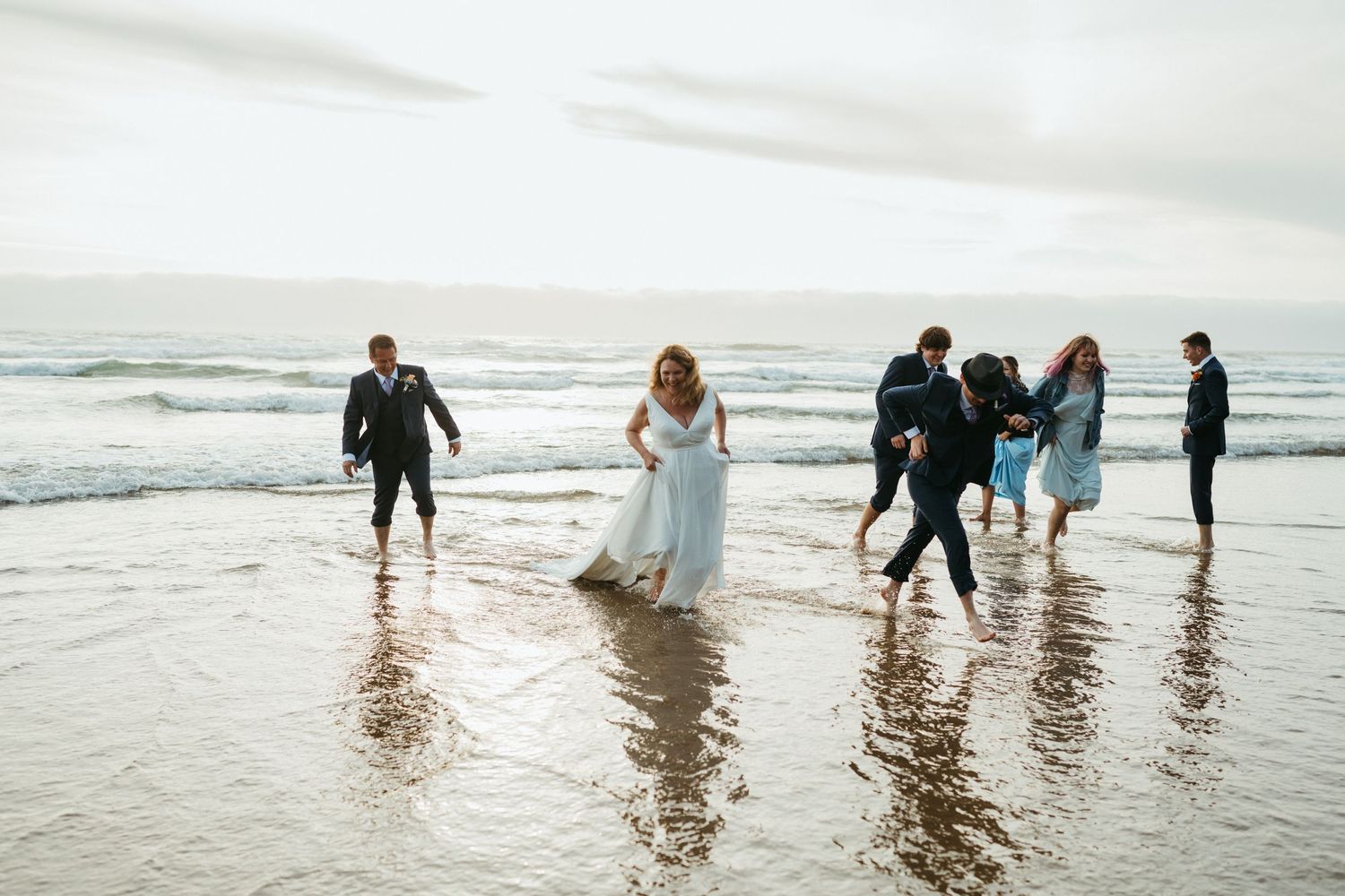 Wedding party members dance and splash in shallow ocean waters during celebration.