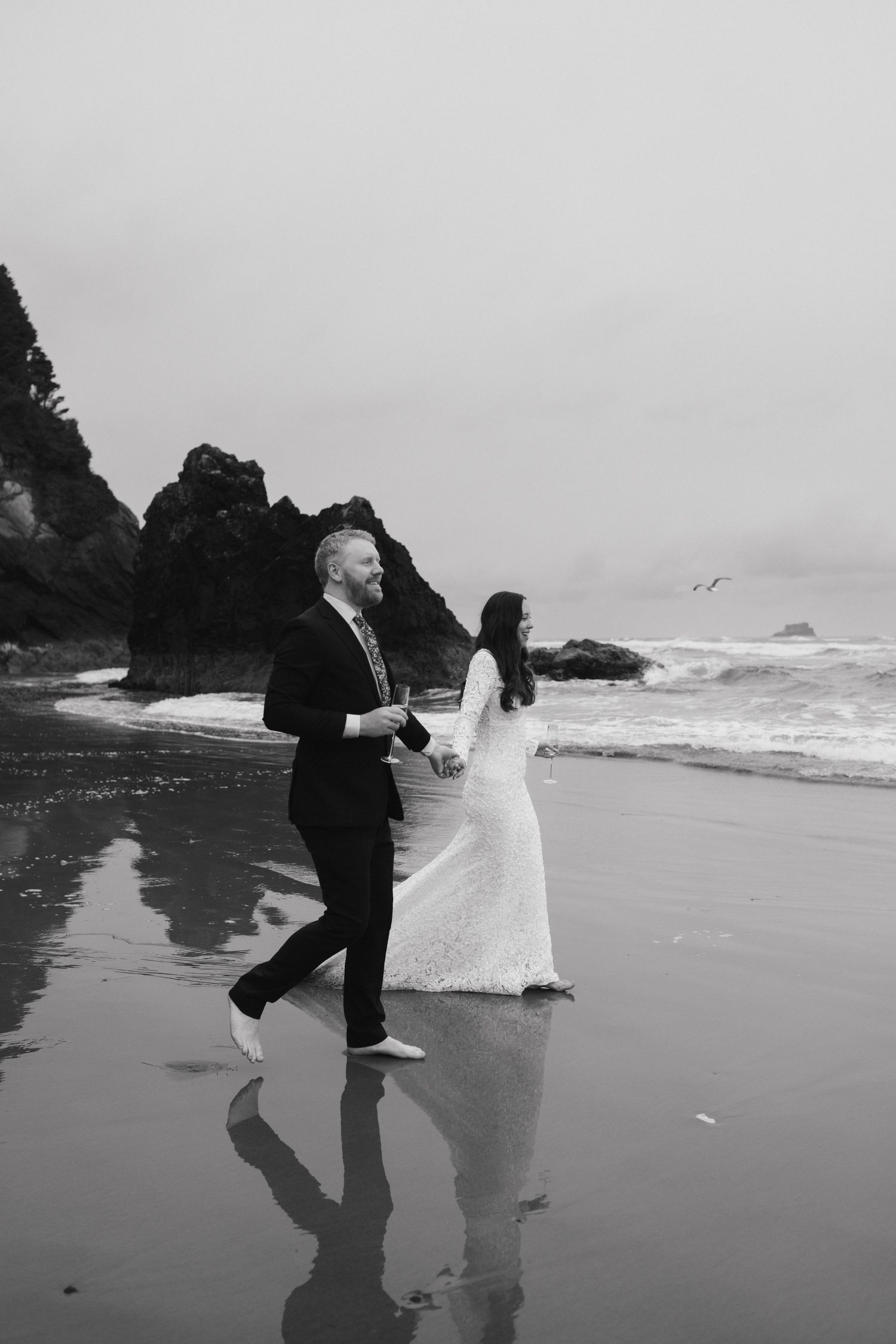 Black and white photo of couple strolling on reflective wet beach with rocky cliffs.