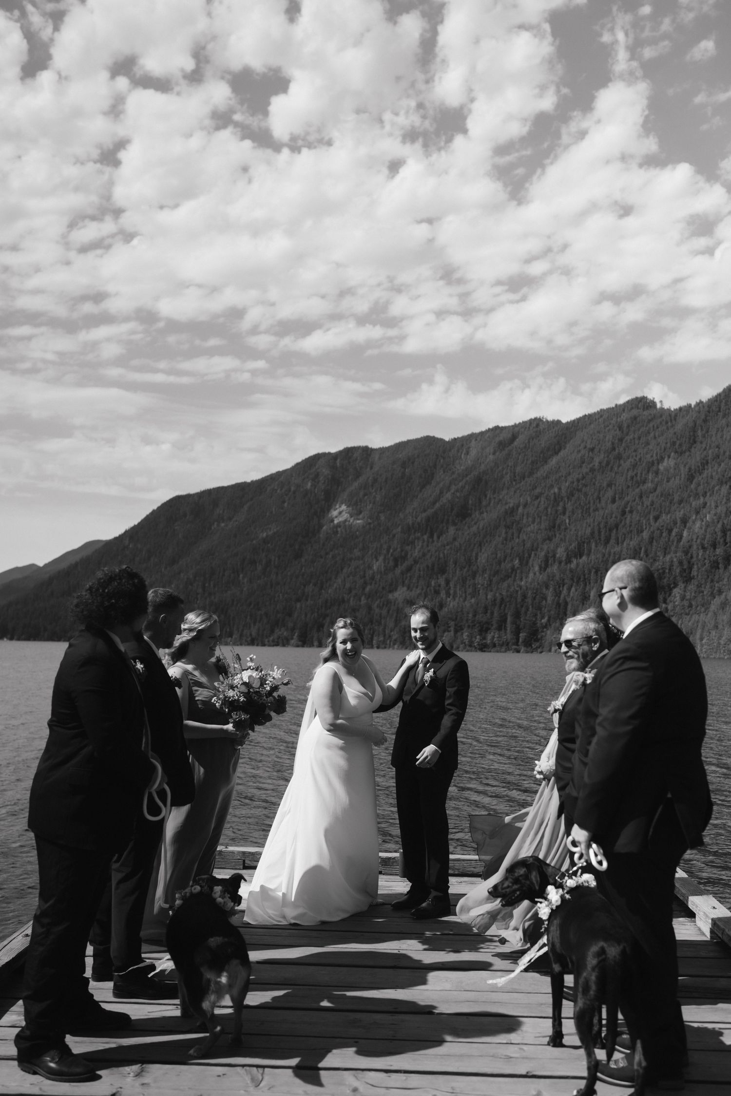Black and white lakeside wedding ceremony with two dogs and mountain backdrop.