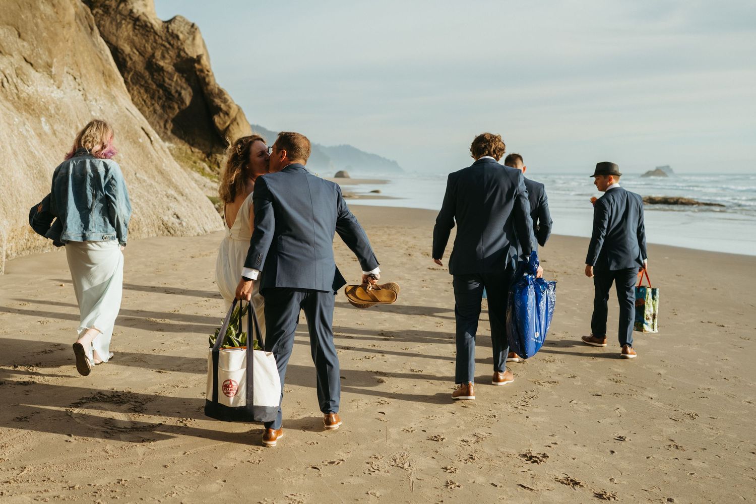 Wedding party members walk along a scenic beach path carrying bags and supplies.