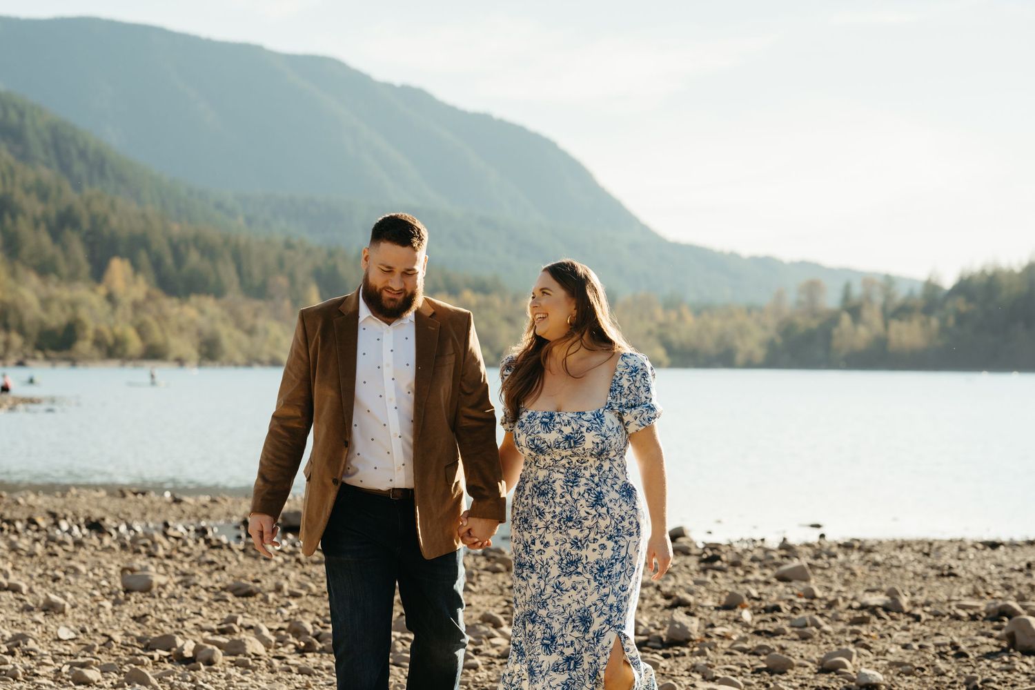 A couple walks hand in hand along a rocky lakeshore with mountains and forest in the background during golden hour.