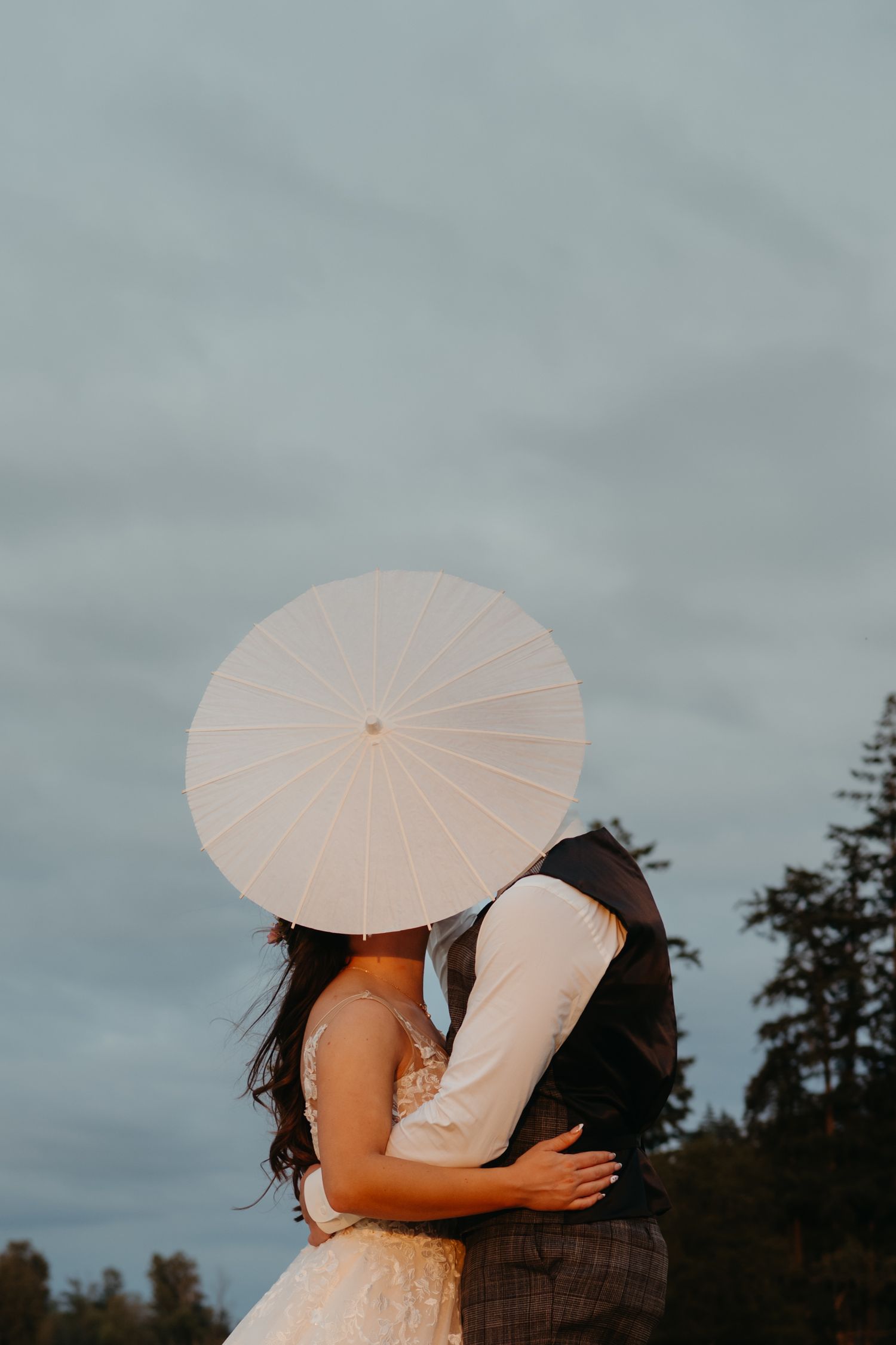 Bride and groom kissing behind a white paper parasol umbrella outdoors on their wedding day.
