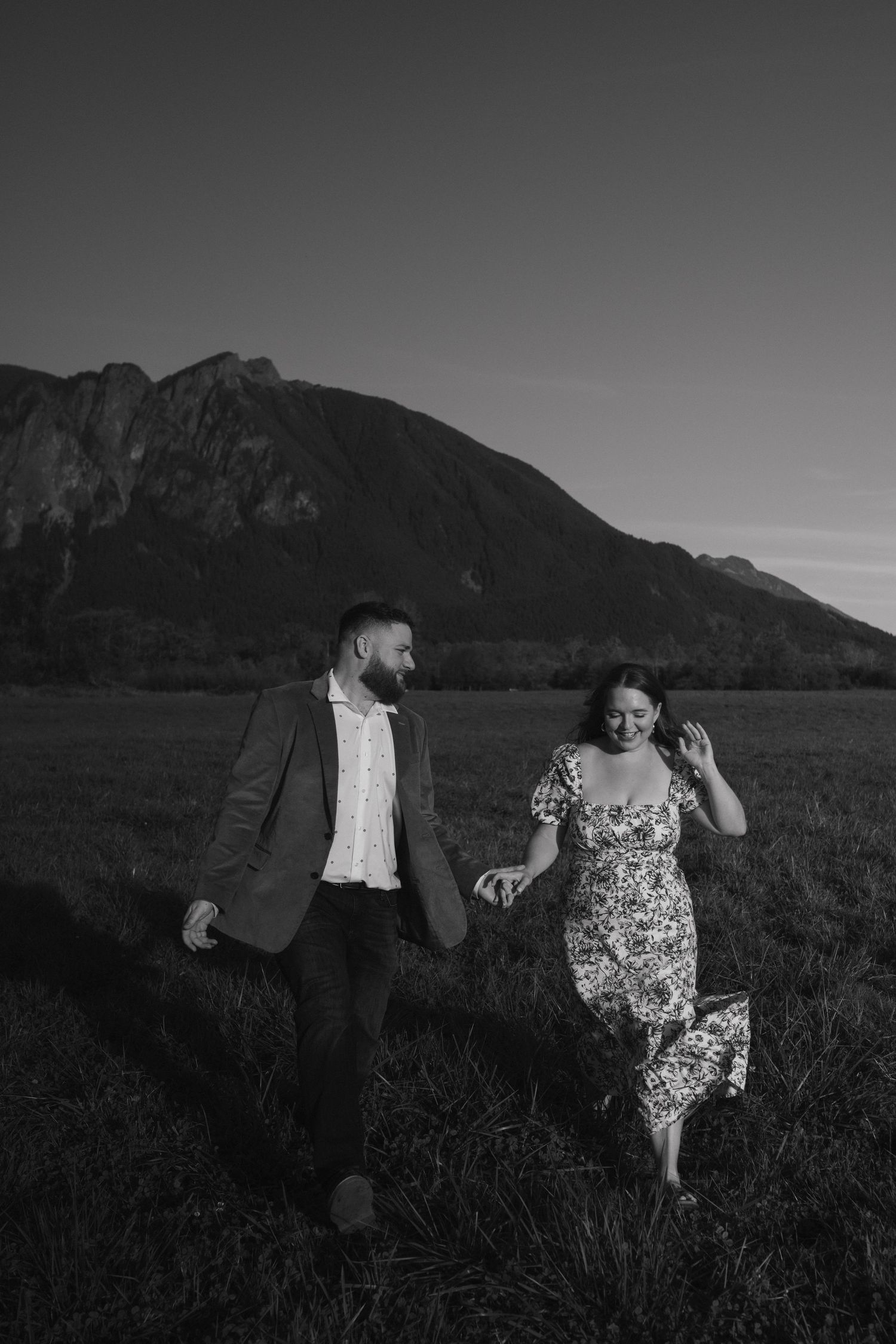 A couple runs playfully through a field with dramatic mountain backdrop at dusk.