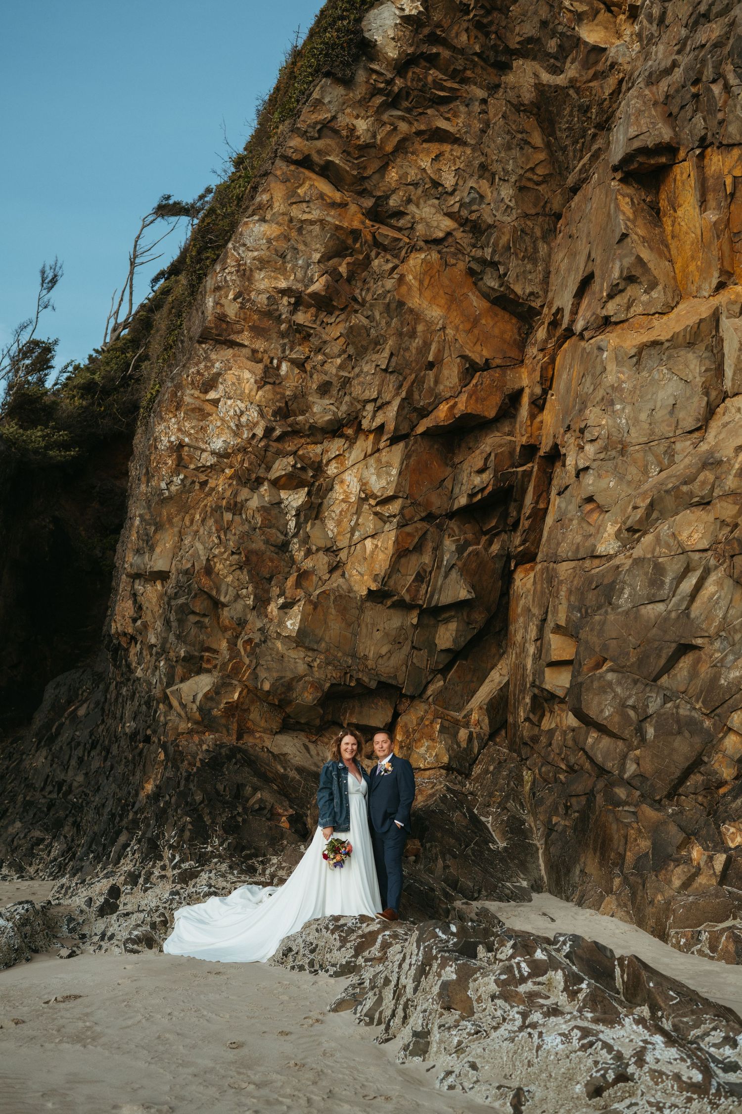 A wedding couple poses against a dramatic rocky cliff face at the beach during sunset.