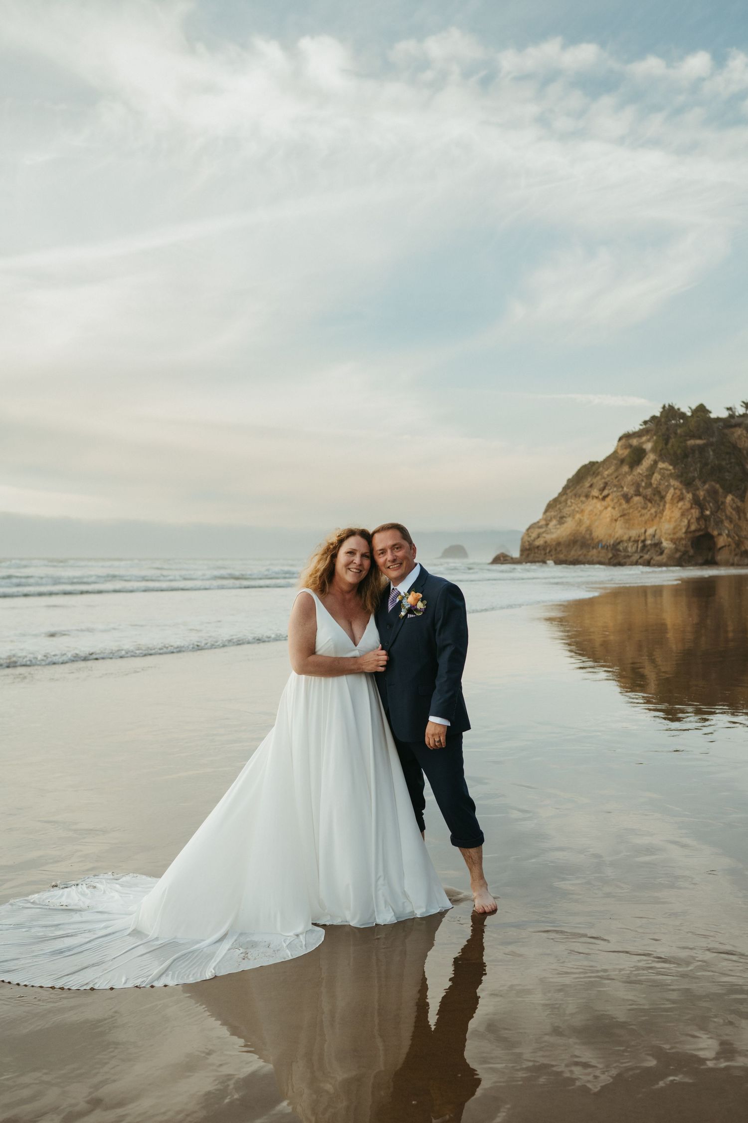 A beach wedding portrait with couple standing in shallow ocean water near coastal cliffs.