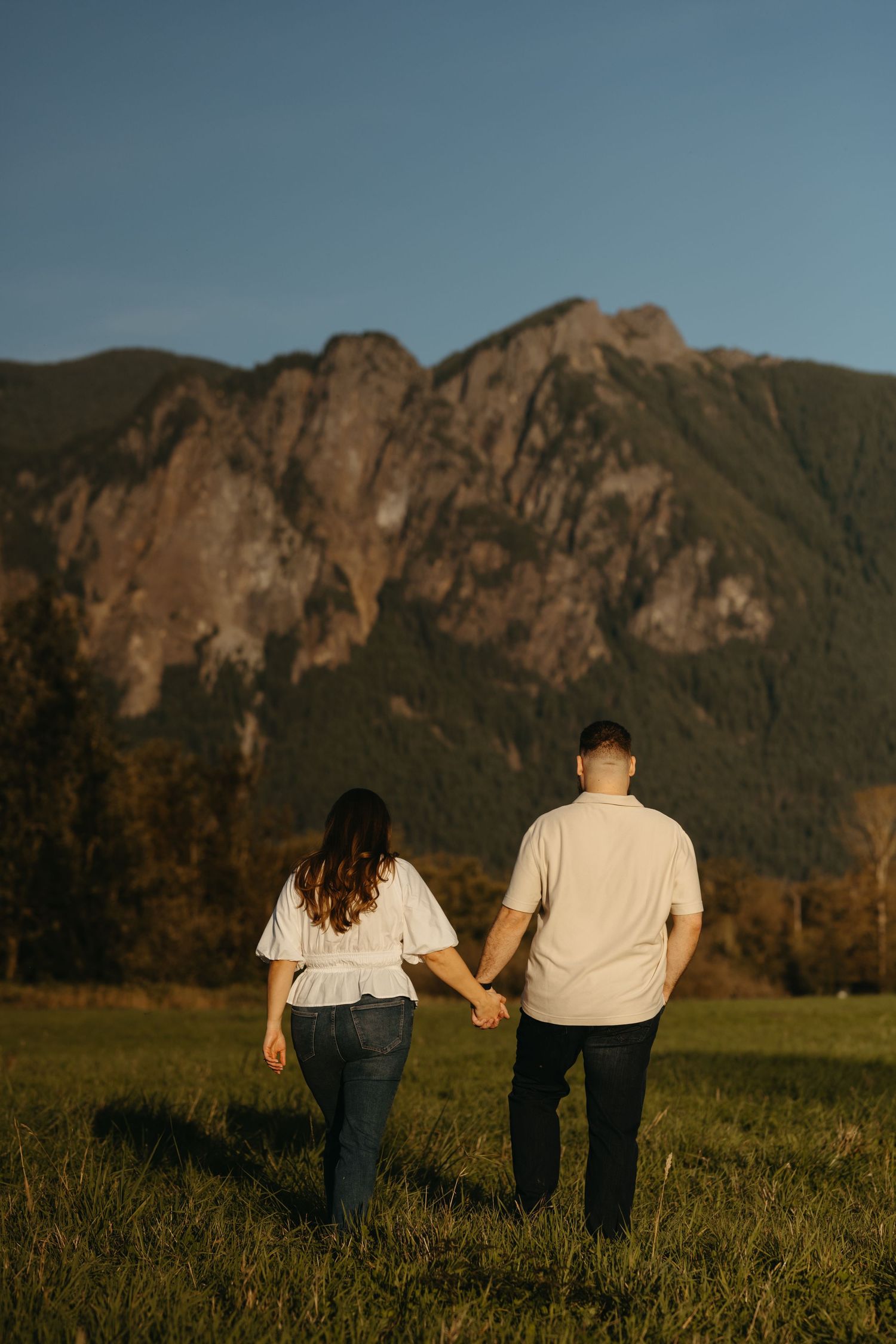 Couple walking hand in hand in a field with dramatic mountain backdrop at sunset.