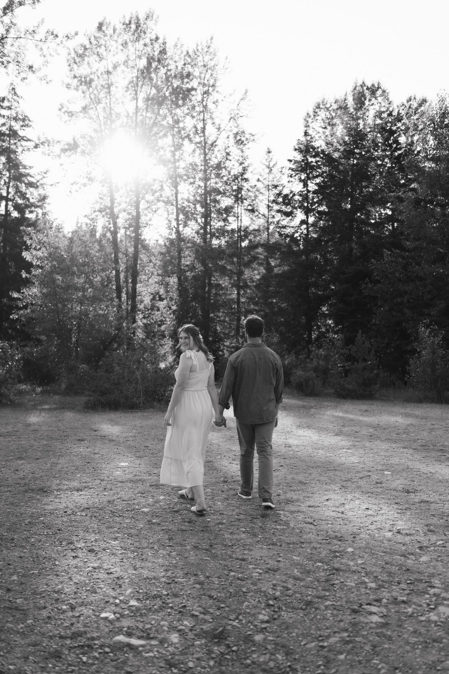 Black and white photo of couple walking away on dirt path with sunburst through trees.