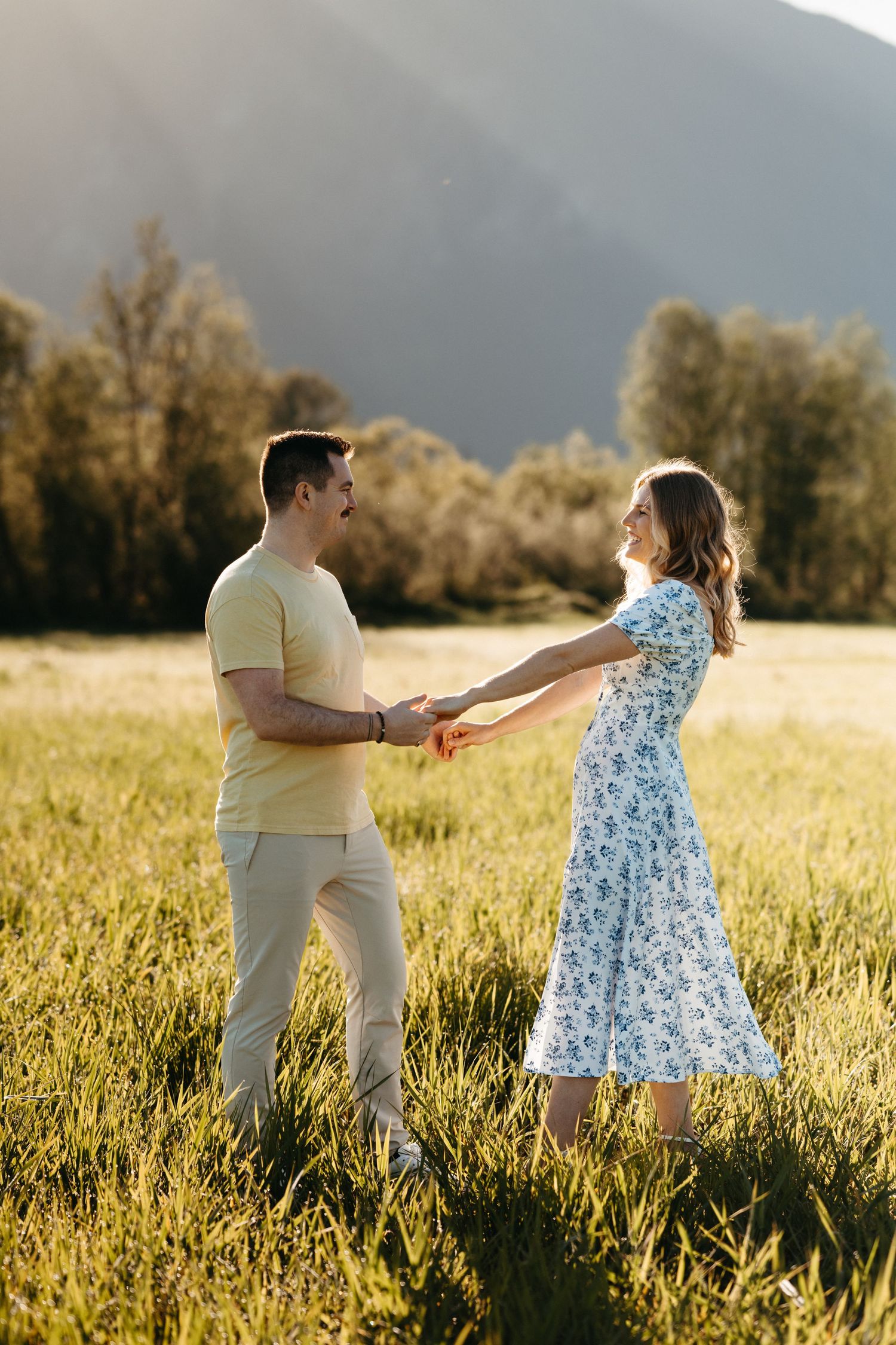 A couple holds hands while dancing in a sunny meadow with mountains in the background.