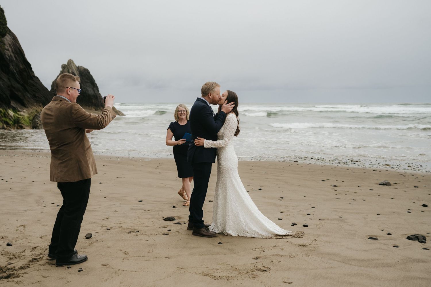 A photographer captures newlyweds sharing their first kiss on a cloudy beach.