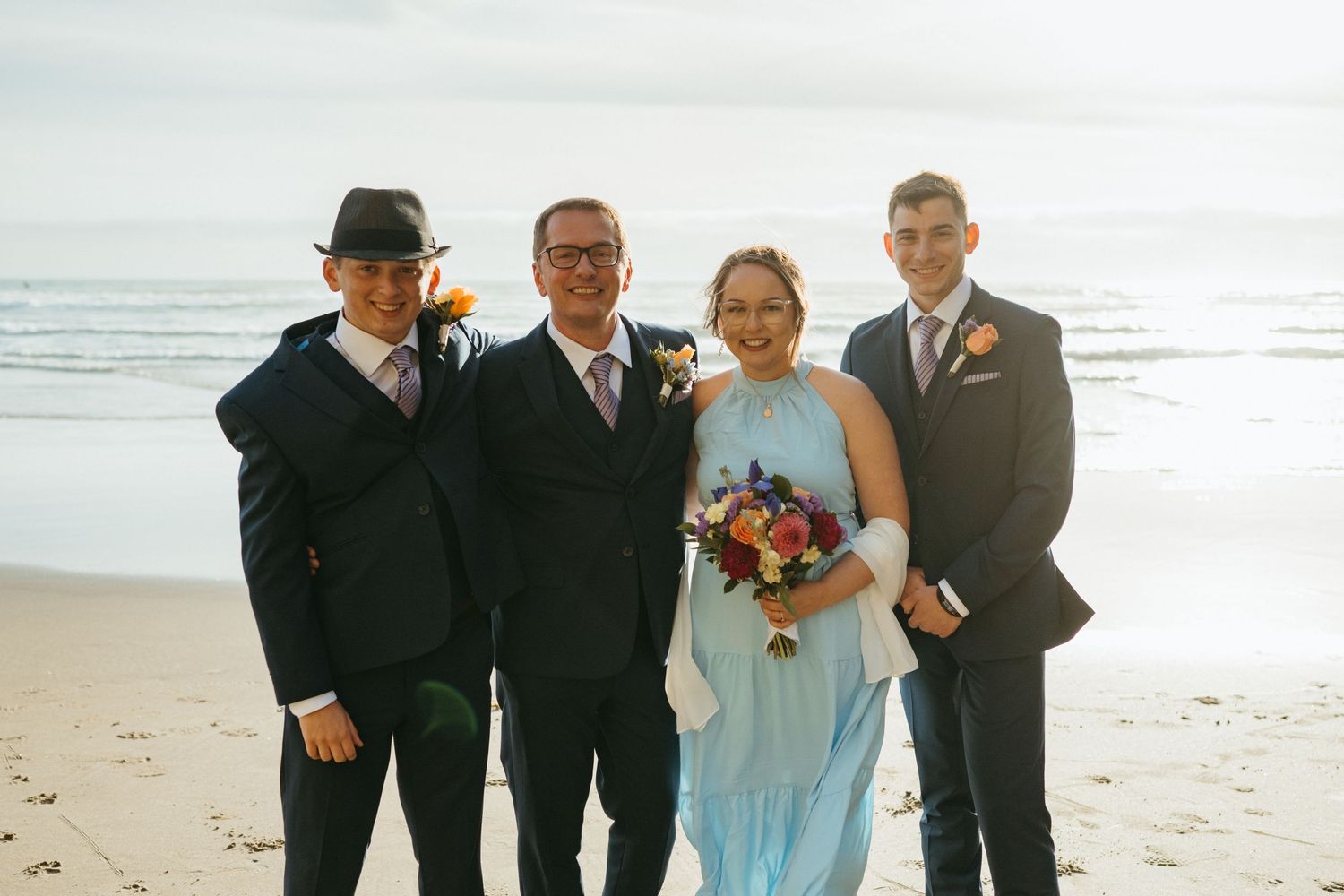 Four wedding attendants in formal attire pose together on a sunny beach.