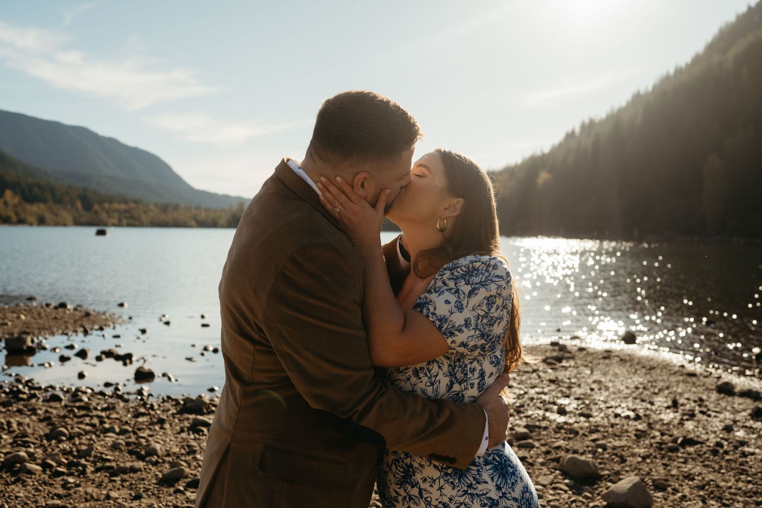 A couple shares an intimate moment on a rocky beach with golden sunlight and mountains in the background.