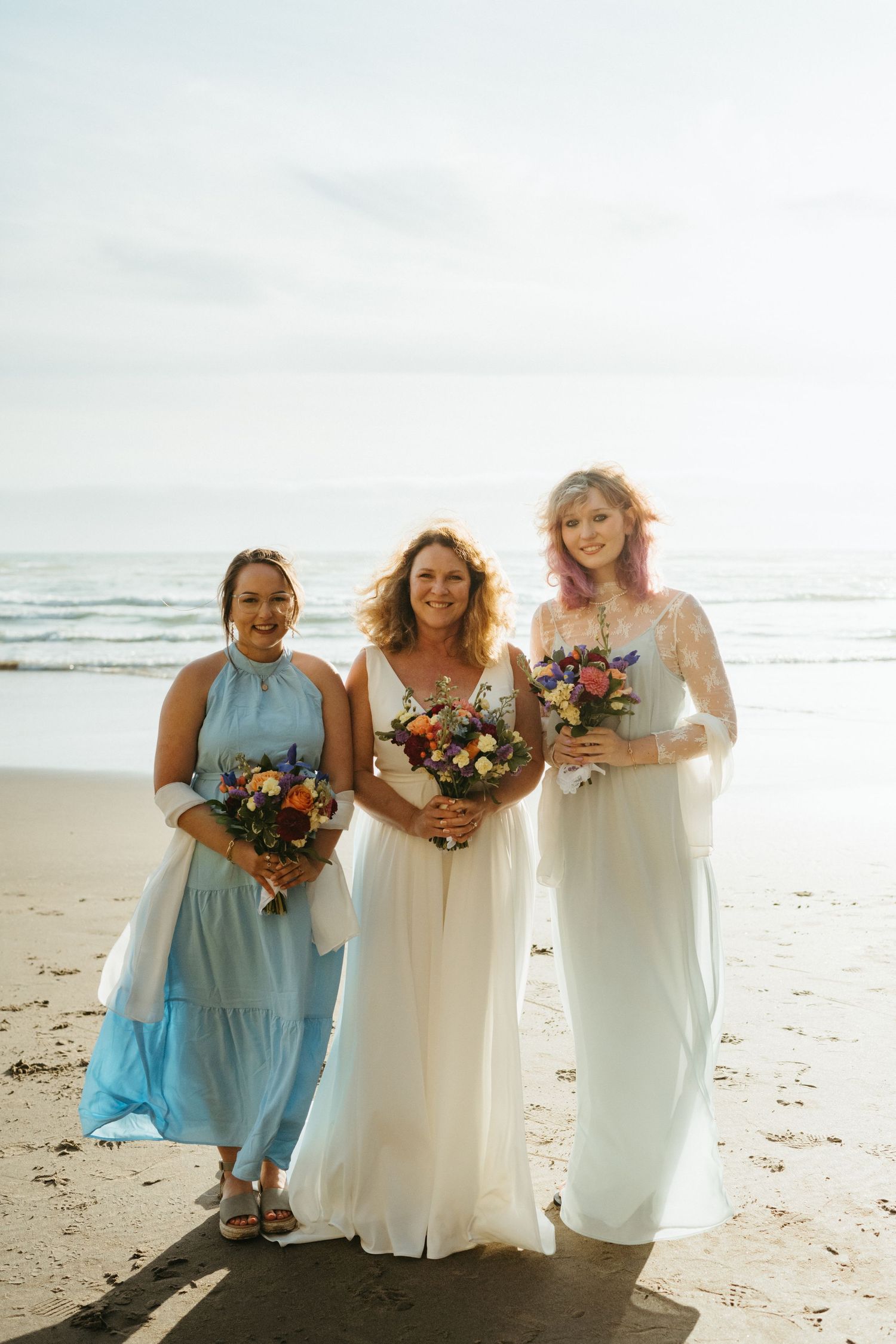 Three attendants in blue and white dresses holding floral bouquets on a beach.