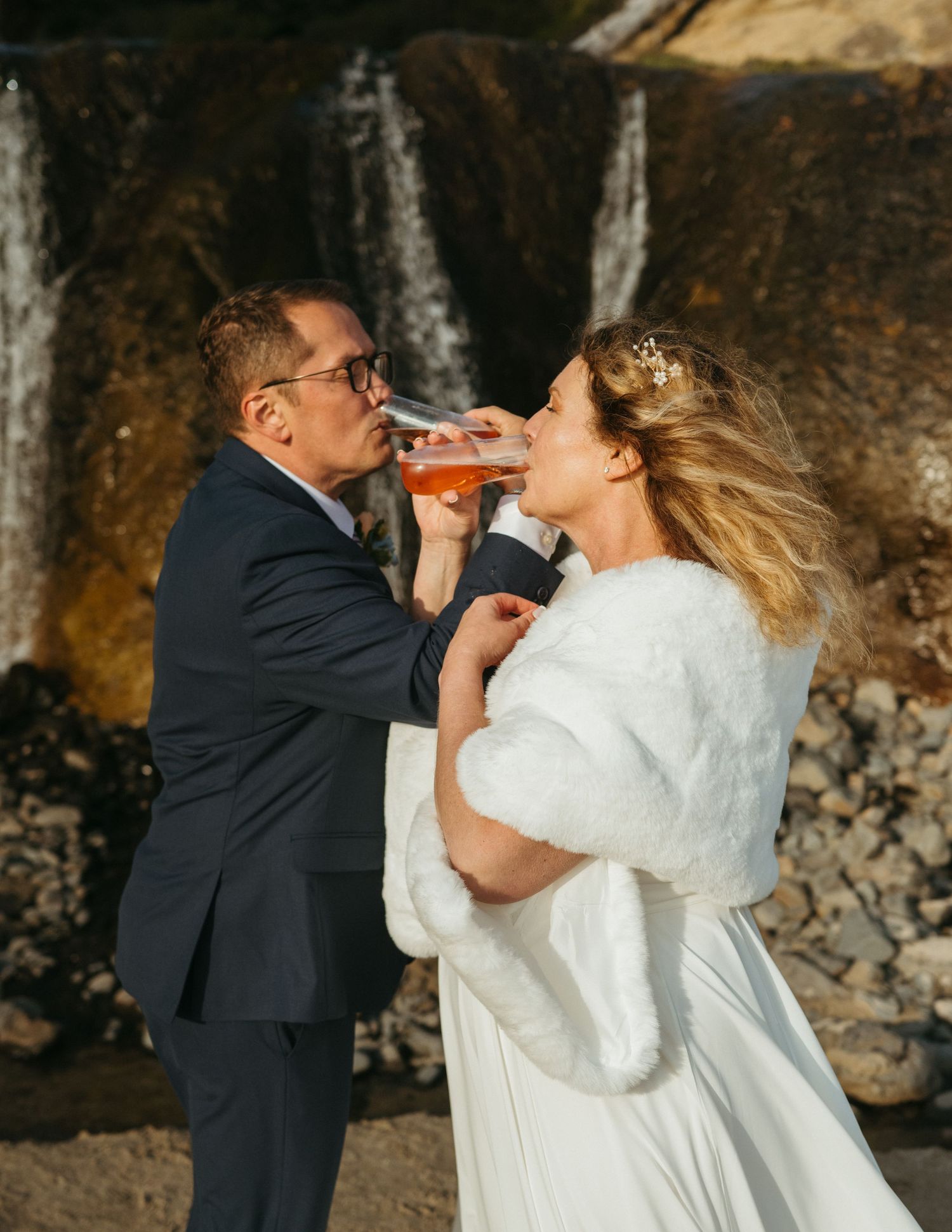 Bride in flowing white dress stands before scenic waterfall on beach.