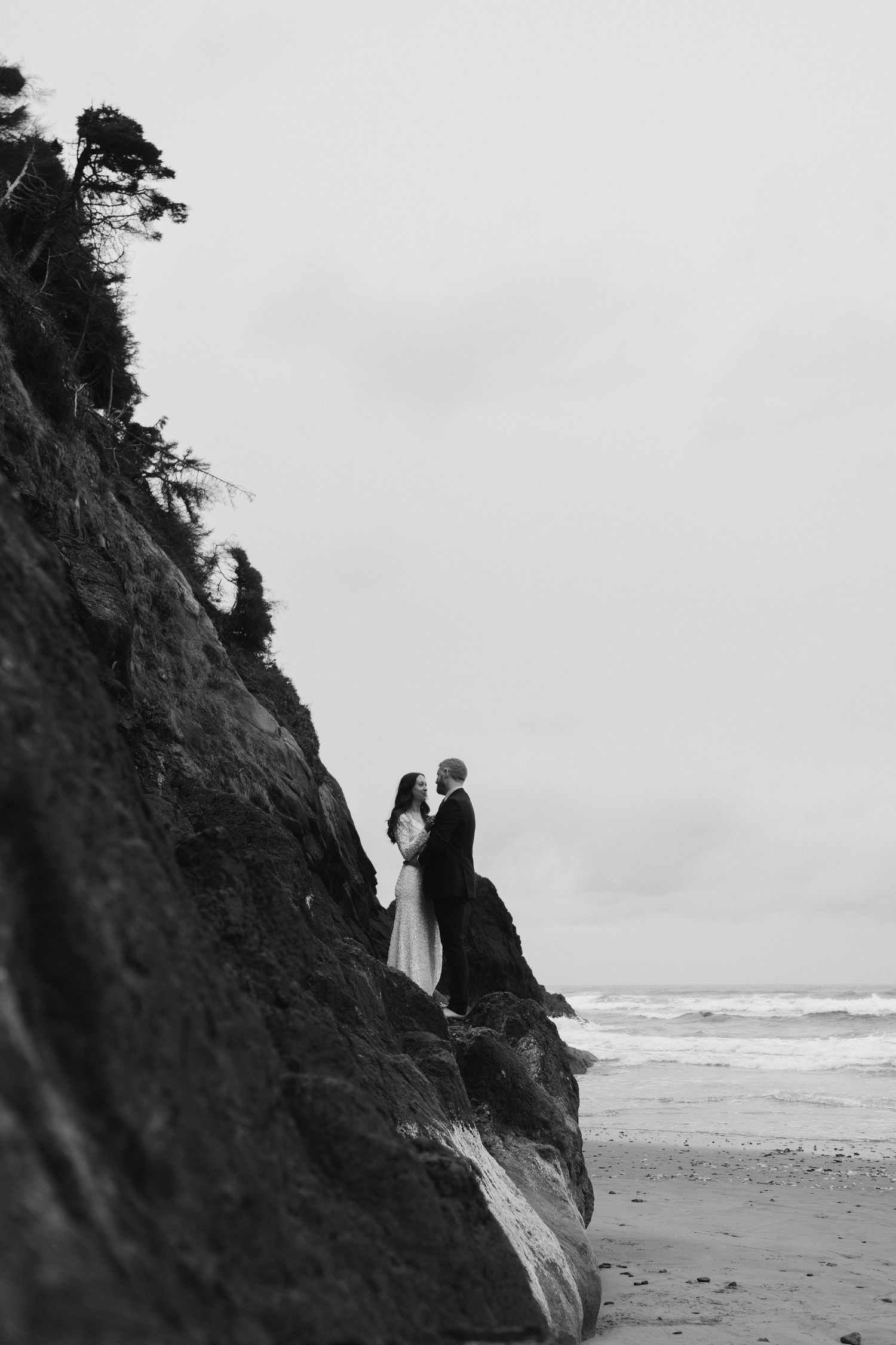 A dramatic black and white photo of newlyweds standing on a coastal cliff overlooking the ocean.