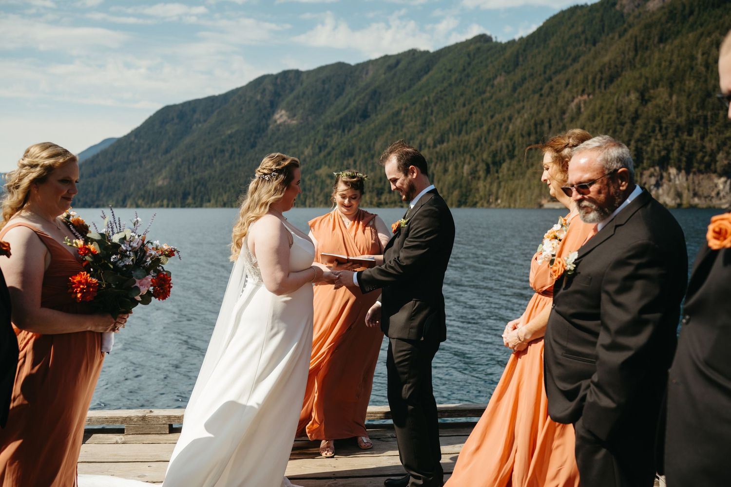 Wedding ceremony taking place on a dock overlooking mountains and a lake with bridesmaids in orange dresses.