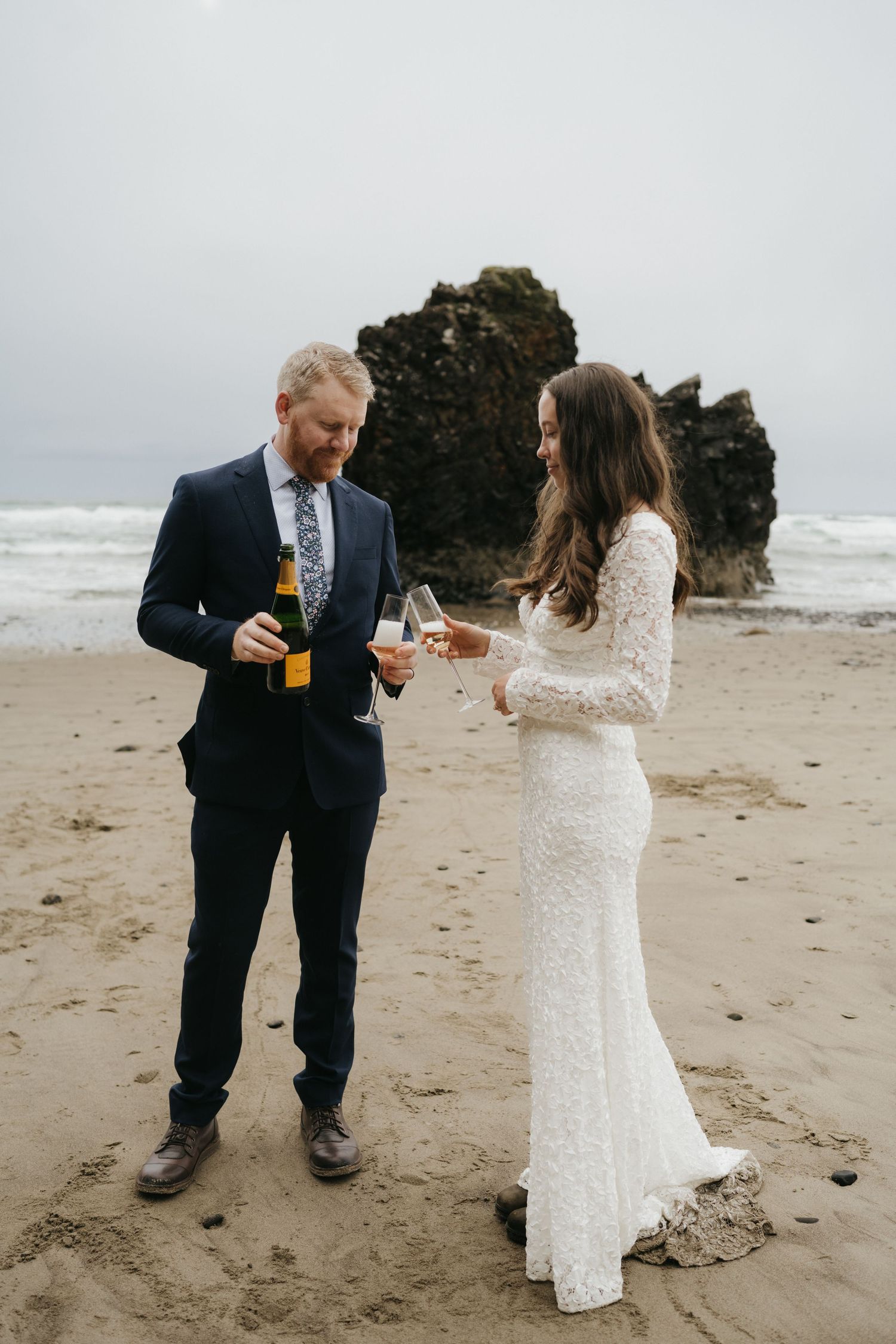 Couple sharing drinks on a beach with rugged coastal rocks in the background.
