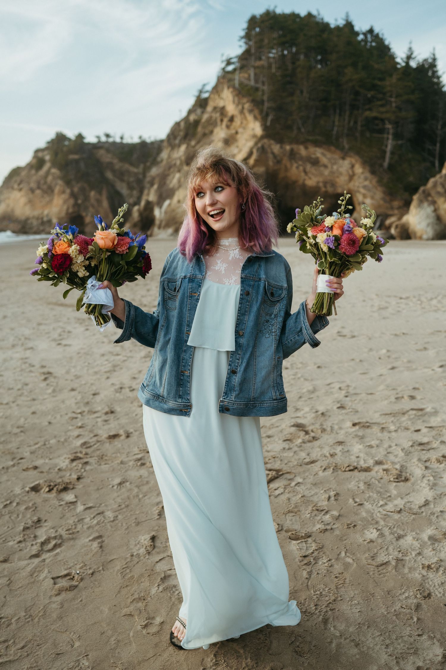 Person in denim jacket and white dress holds two vibrant floral bouquets on a beach.