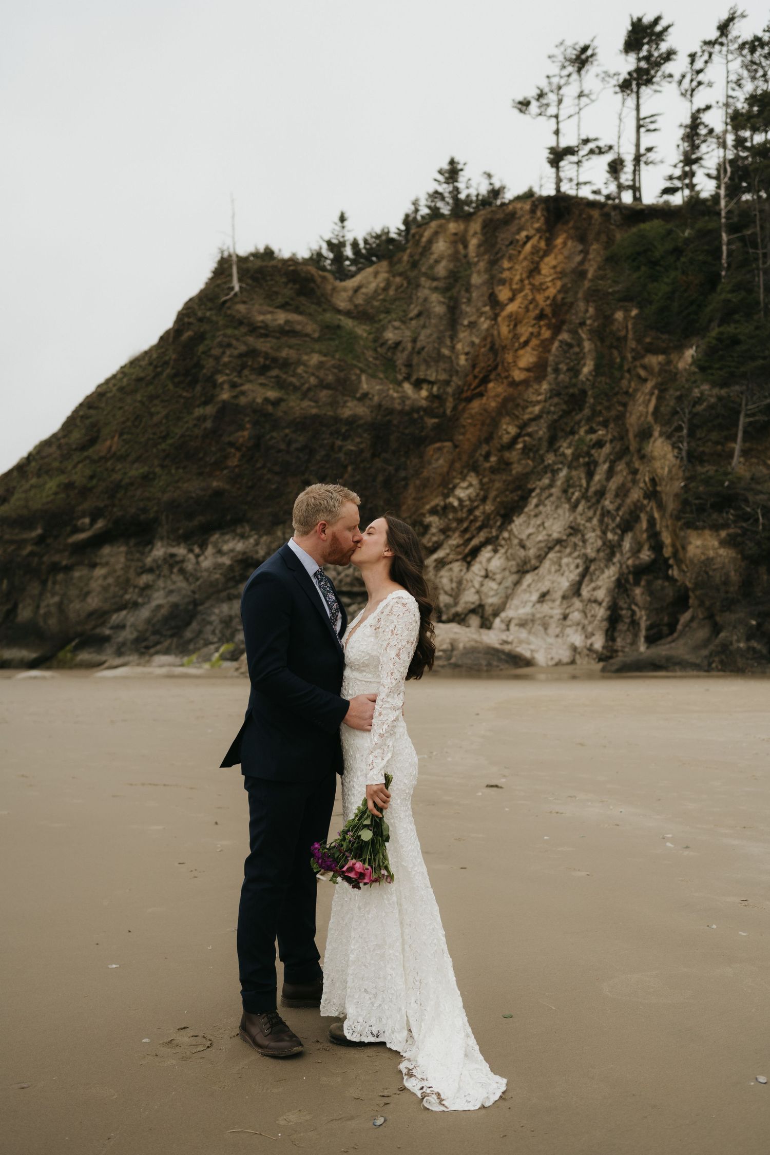 Newlyweds sharing an intimate moment on beach with dramatic coastal cliffs behind them.