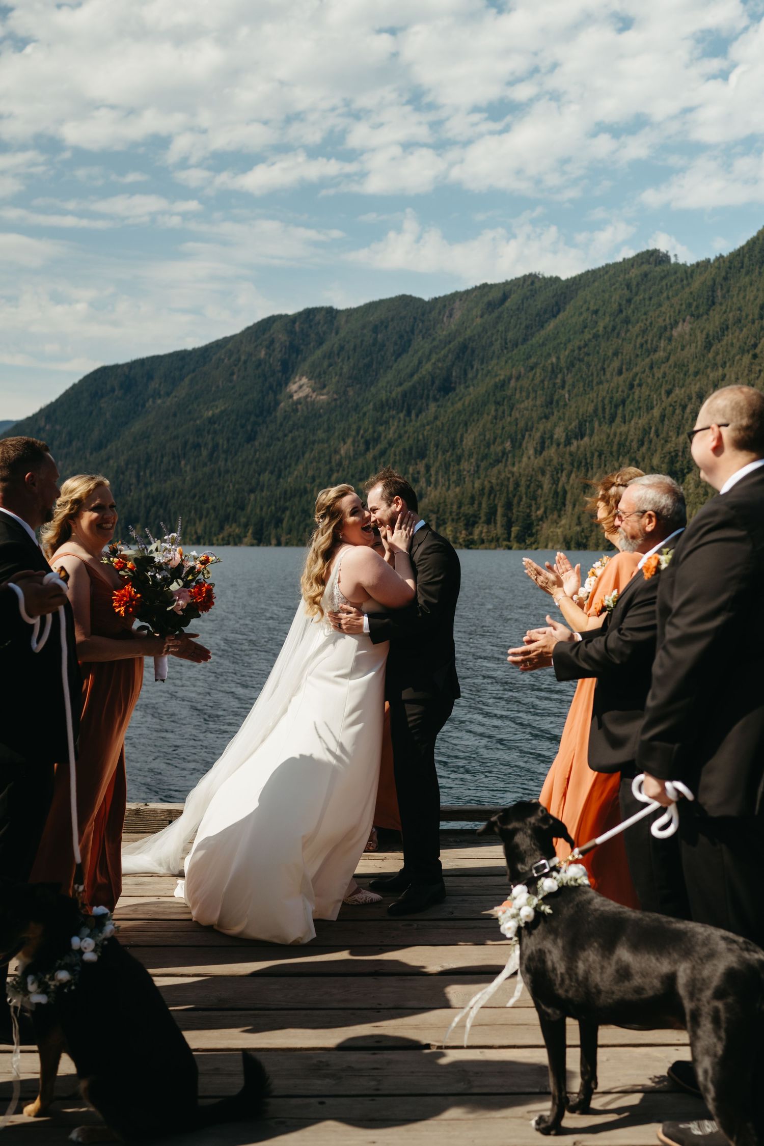 Intimate waterfront wedding ceremony with guests in orange dresses and a black dog watching from the dock.