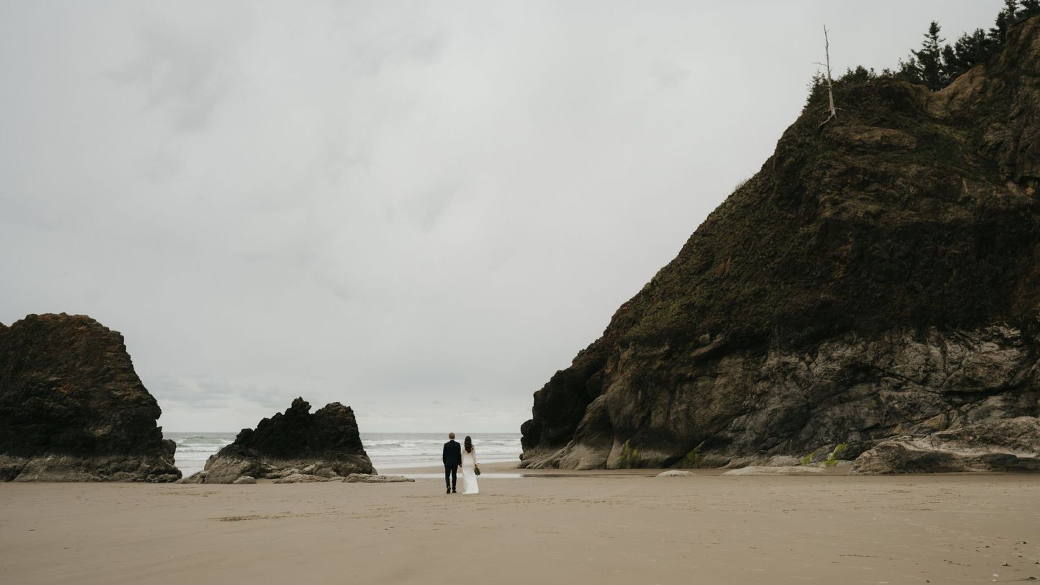 Wedding couple standing small against majestic coastal cliffs on secluded beach.