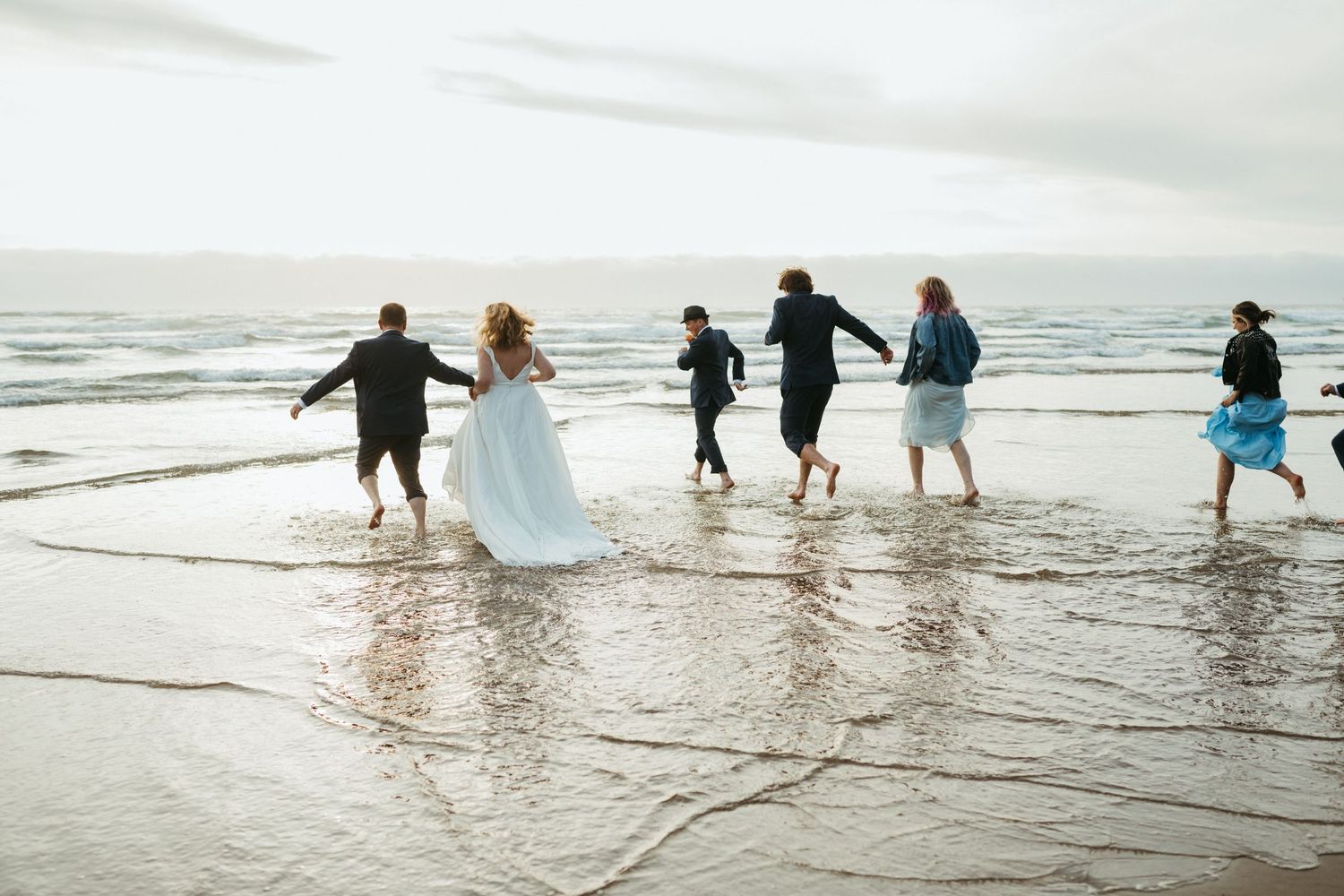 Wedding party splashes and plays in the ocean waves during beach celebration.