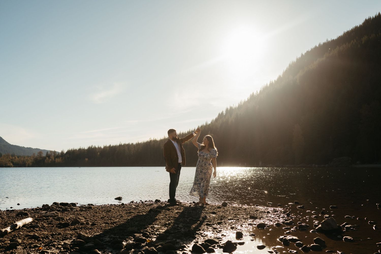 A silhouetted couple stands together on a rocky beach during sunset with mountains in the background.