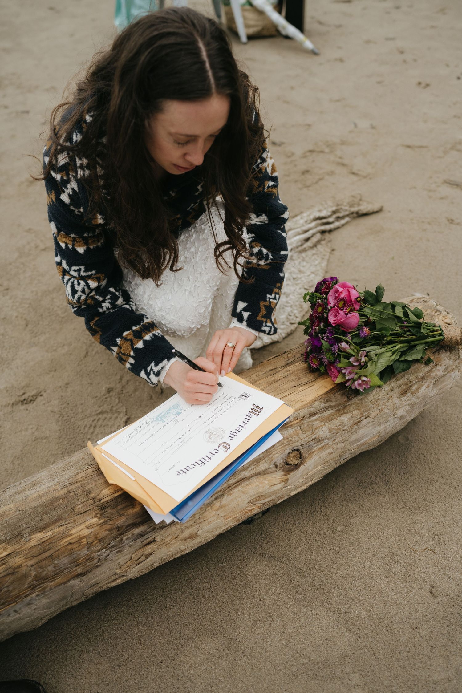 A person in a floral jacket signs documents on driftwood with pink flowers nearby.