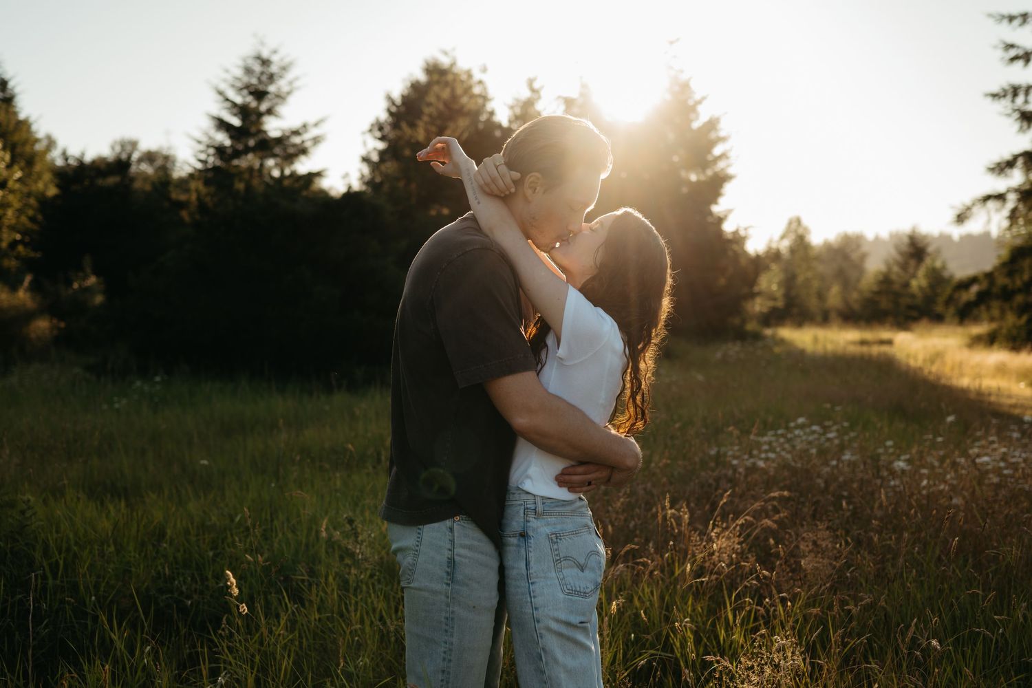 Couple sharing a romantic kiss in a sunlit meadow surrounded by lush green trees during golden hour.