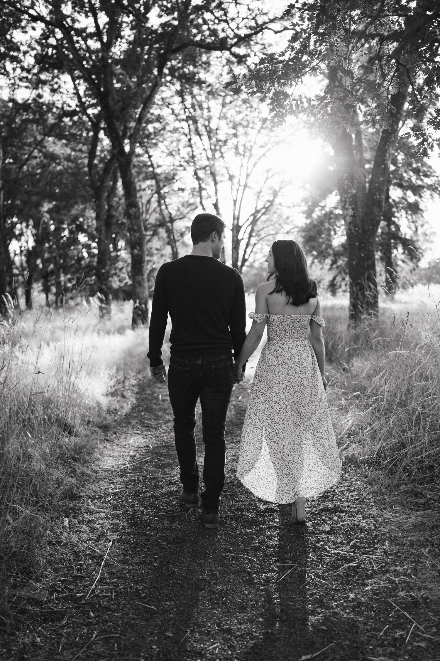 Black and white photo of a couple walking through a tree-lined path with sunlight streaming through.