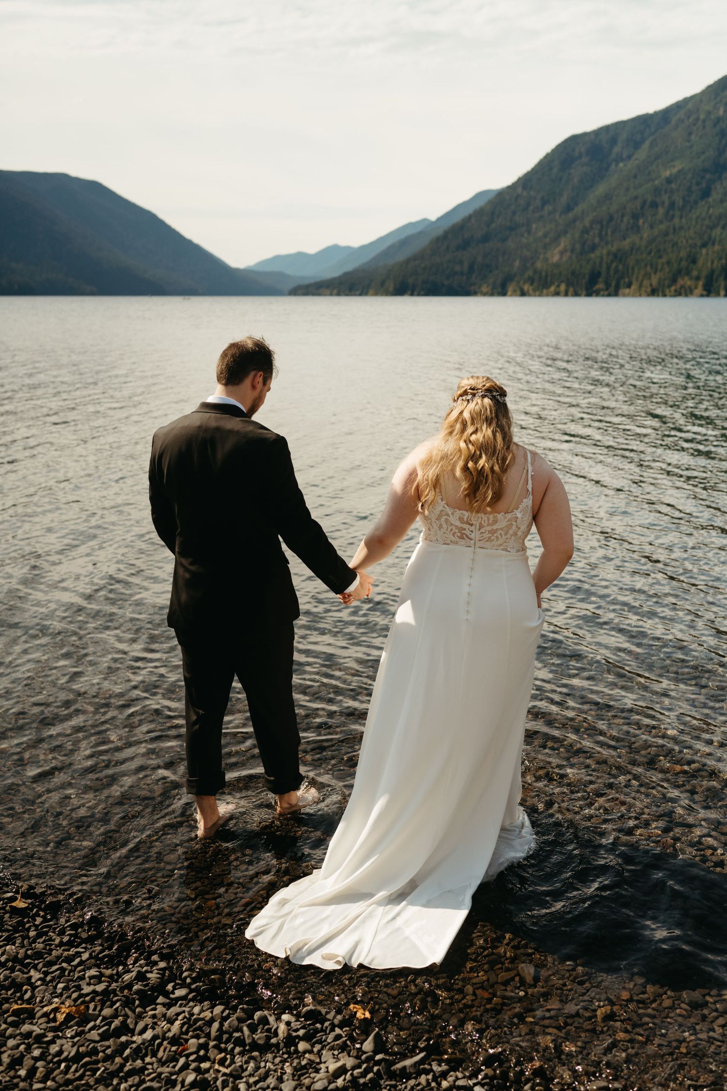 Couple wading in lake water during their wedding photoshoot with mountains in background.