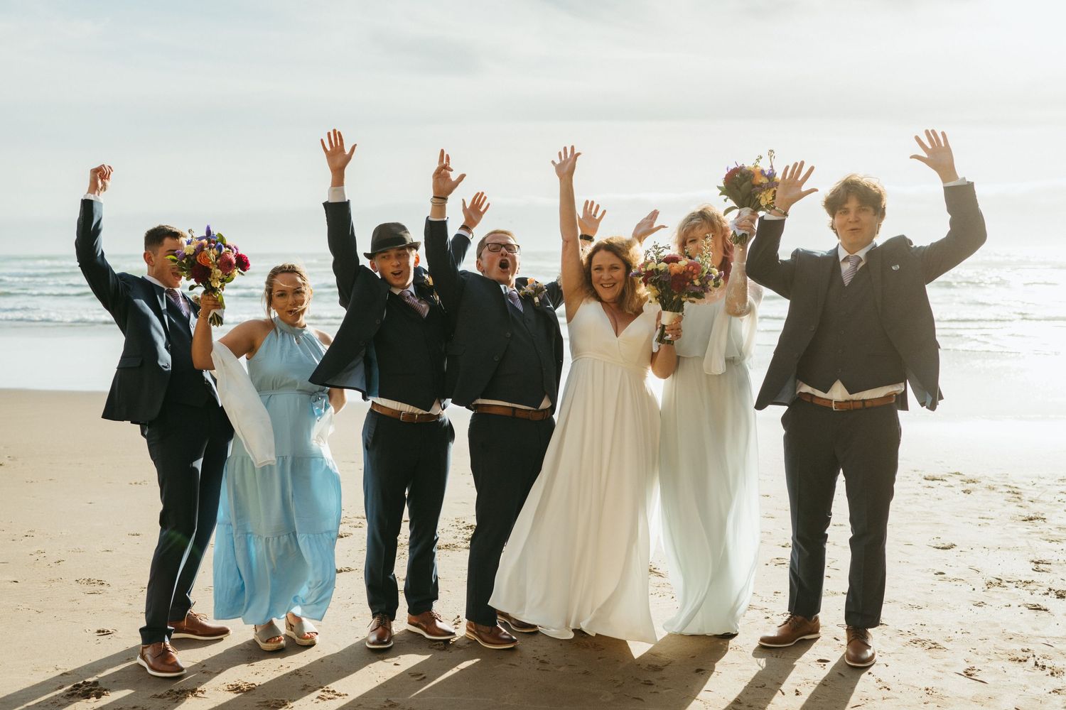 Wedding party celebrating with raised arms on a beach during golden hour.