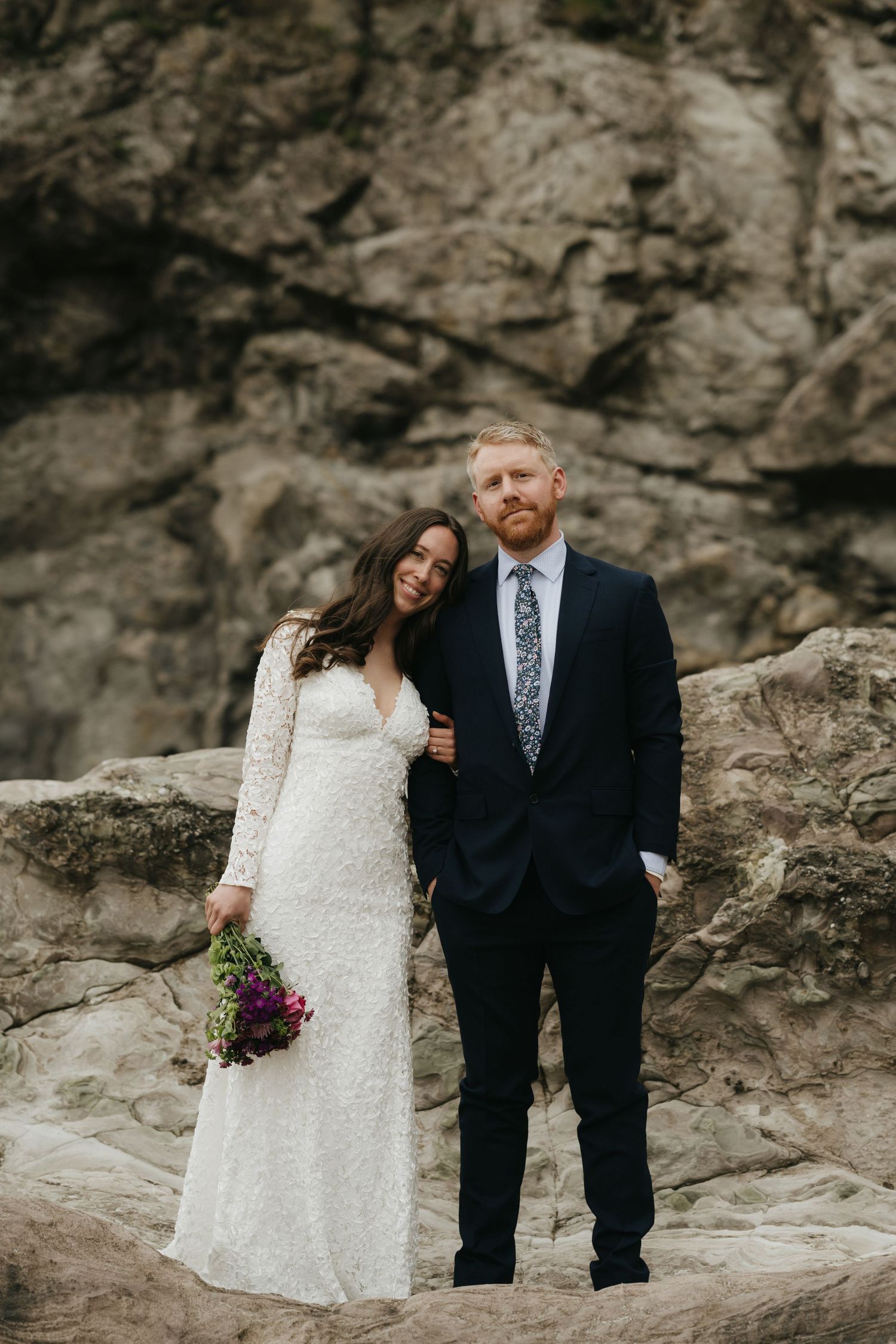 Newlyweds posing against rocky cliff backdrop with bride holding purple bouquet.