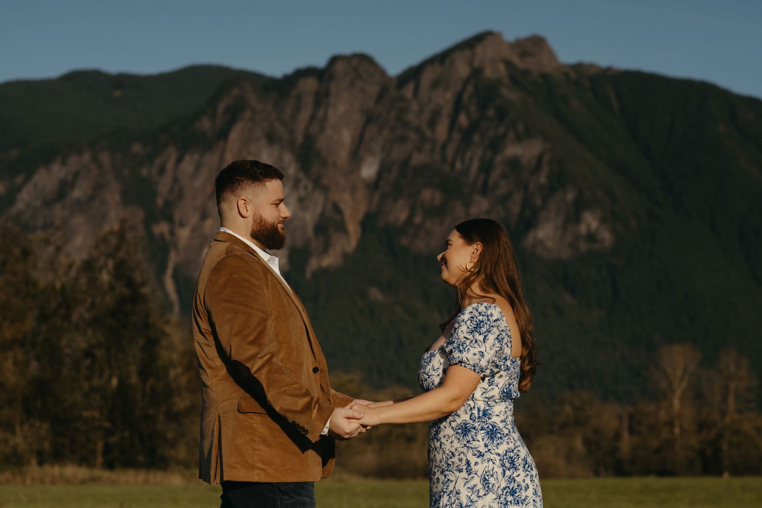Couple holds hands against dramatic mountain backdrop during golden hour sunset engagement photo session.