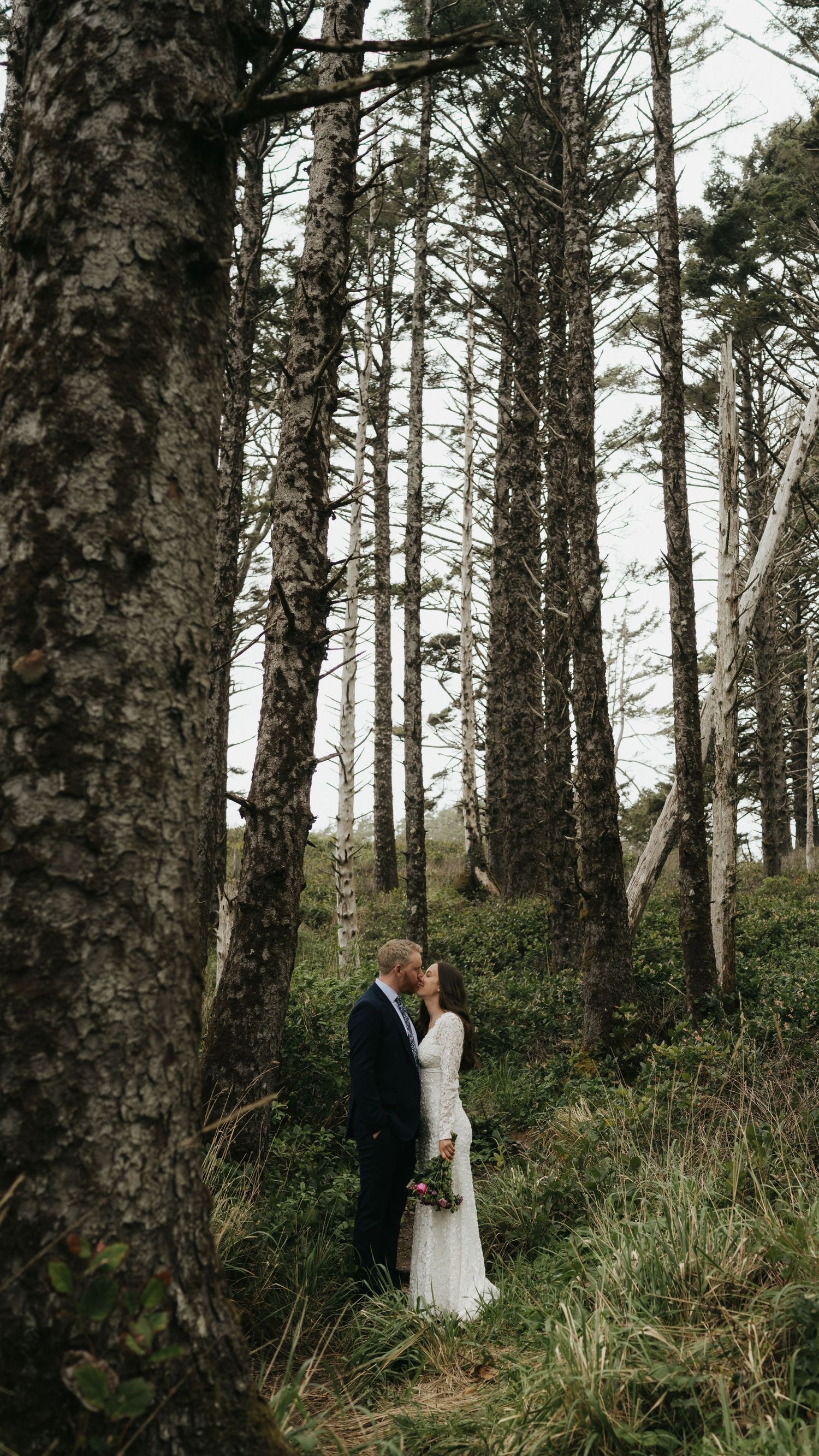 A wedding couple shares an intimate moment surrounded by towering pine trees in a forest.