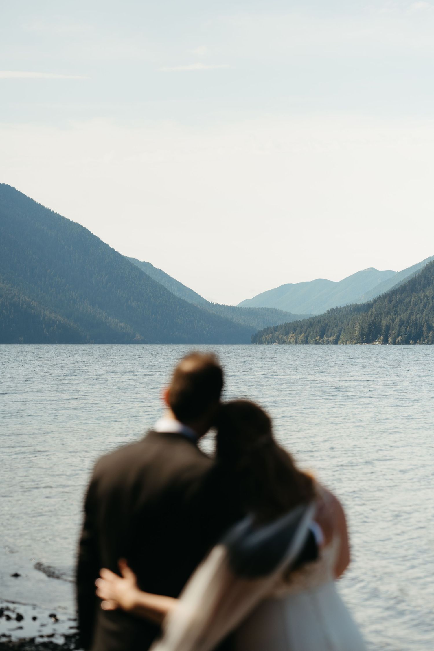 Silhouetted couple embrace while looking out over a peaceful mountain lake landscape.