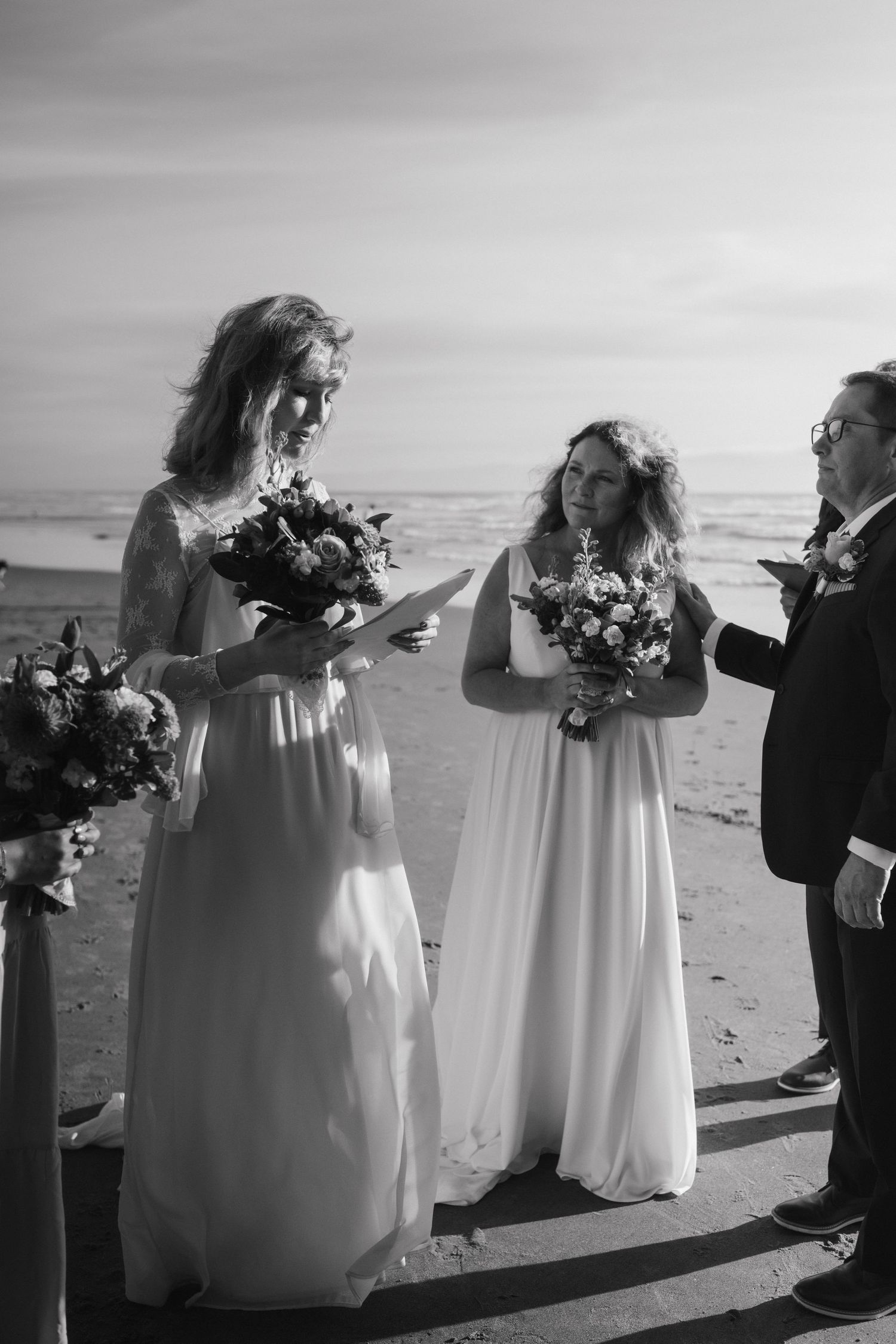 Black and white photo of wedding party with bouquets on beach during ceremony.