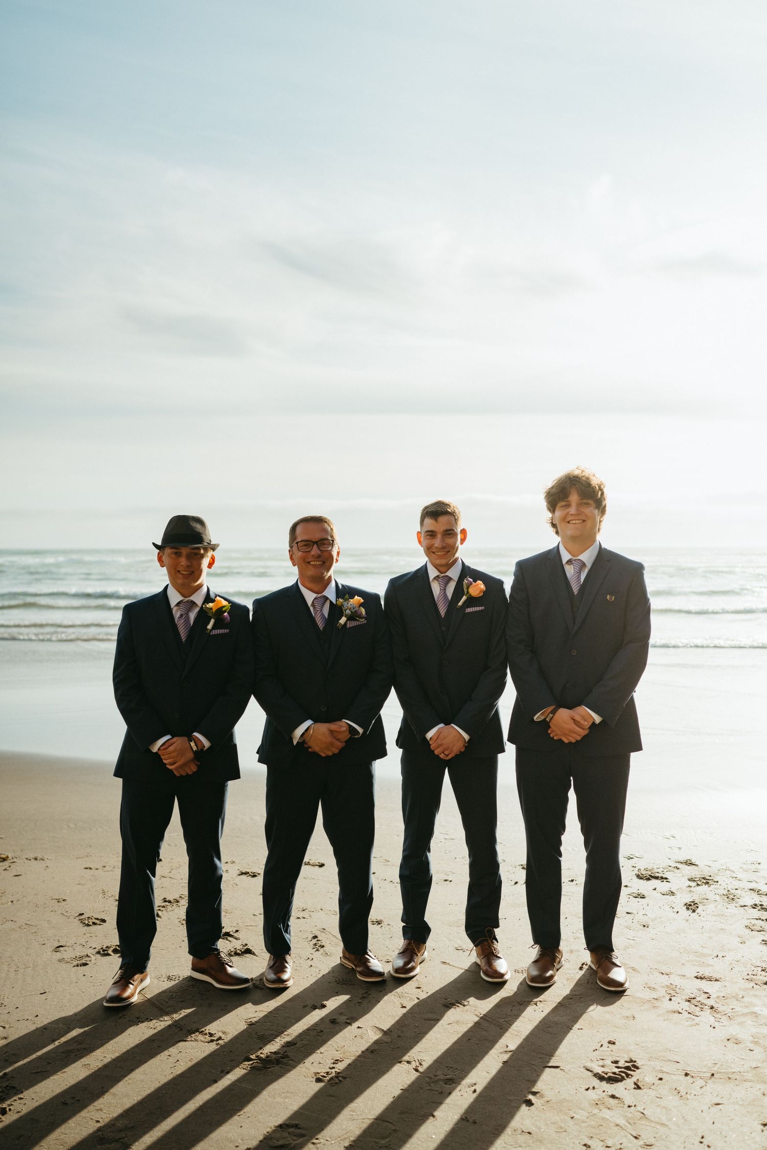 Four groomsmen in dark suits stand on a beach with the ocean behind them casting long shadows on the sand.