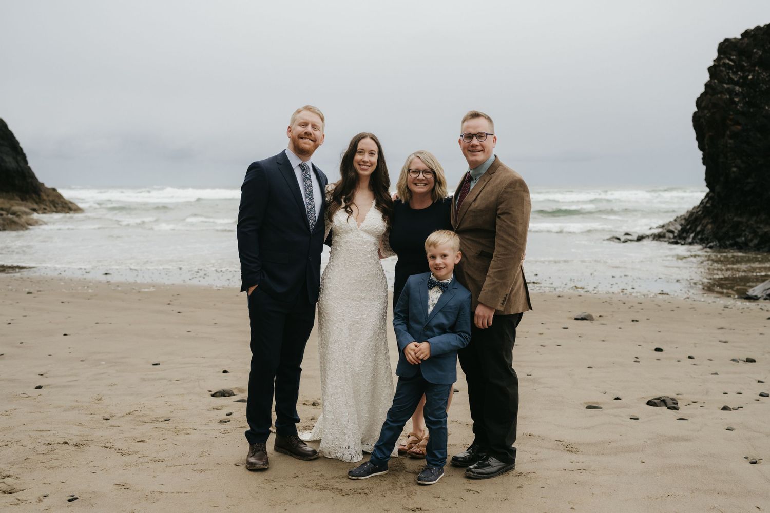 A small wedding group poses for a formal portrait on a moody beach with rocky cliffs.