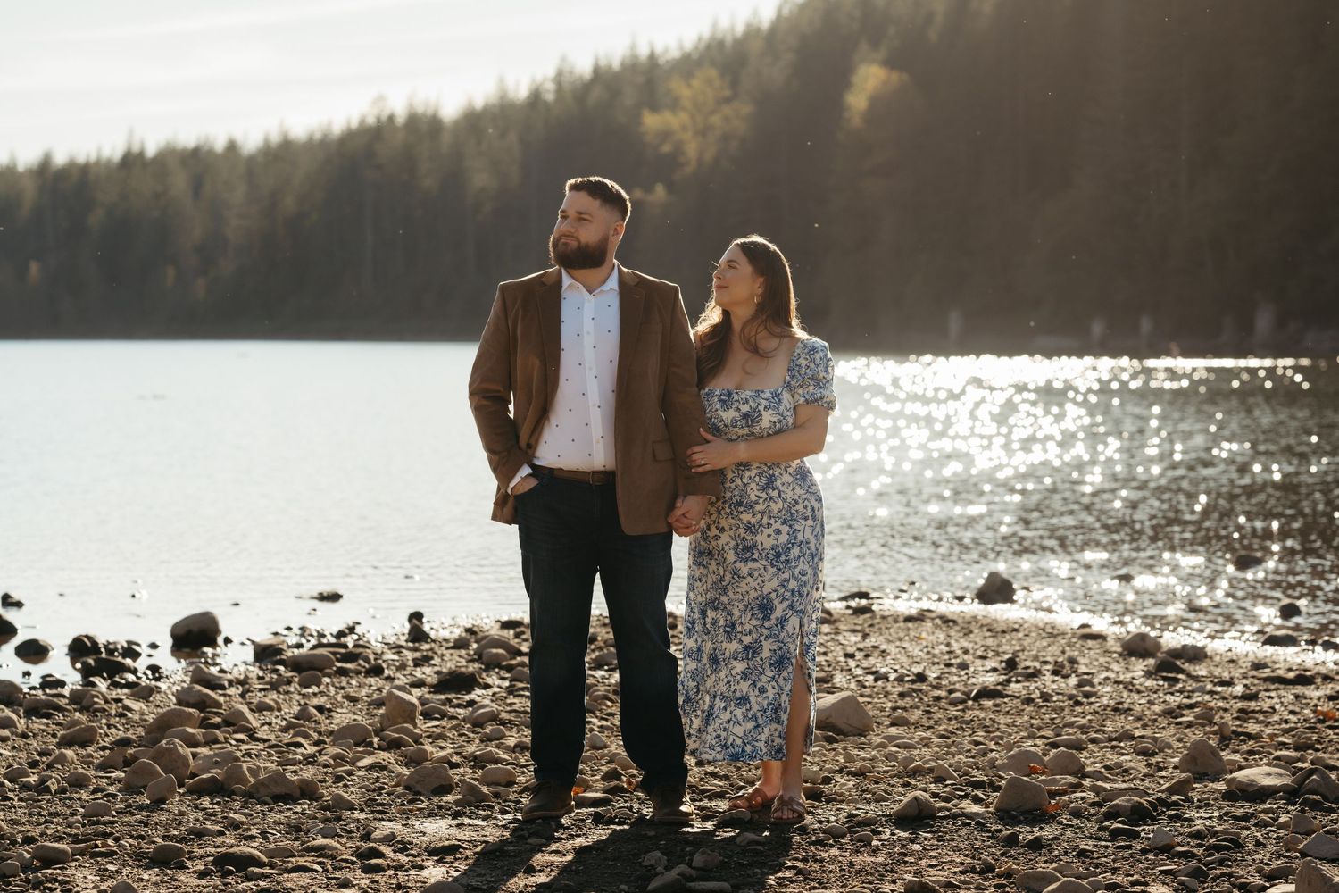 A couple enjoys a sunny moment together on a rocky beach with sparkling water and mountains behind them.