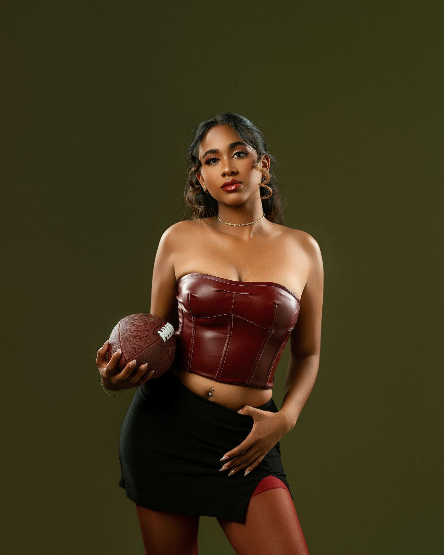 Young woman in burgundy leather corset top holds football against dark olive background.