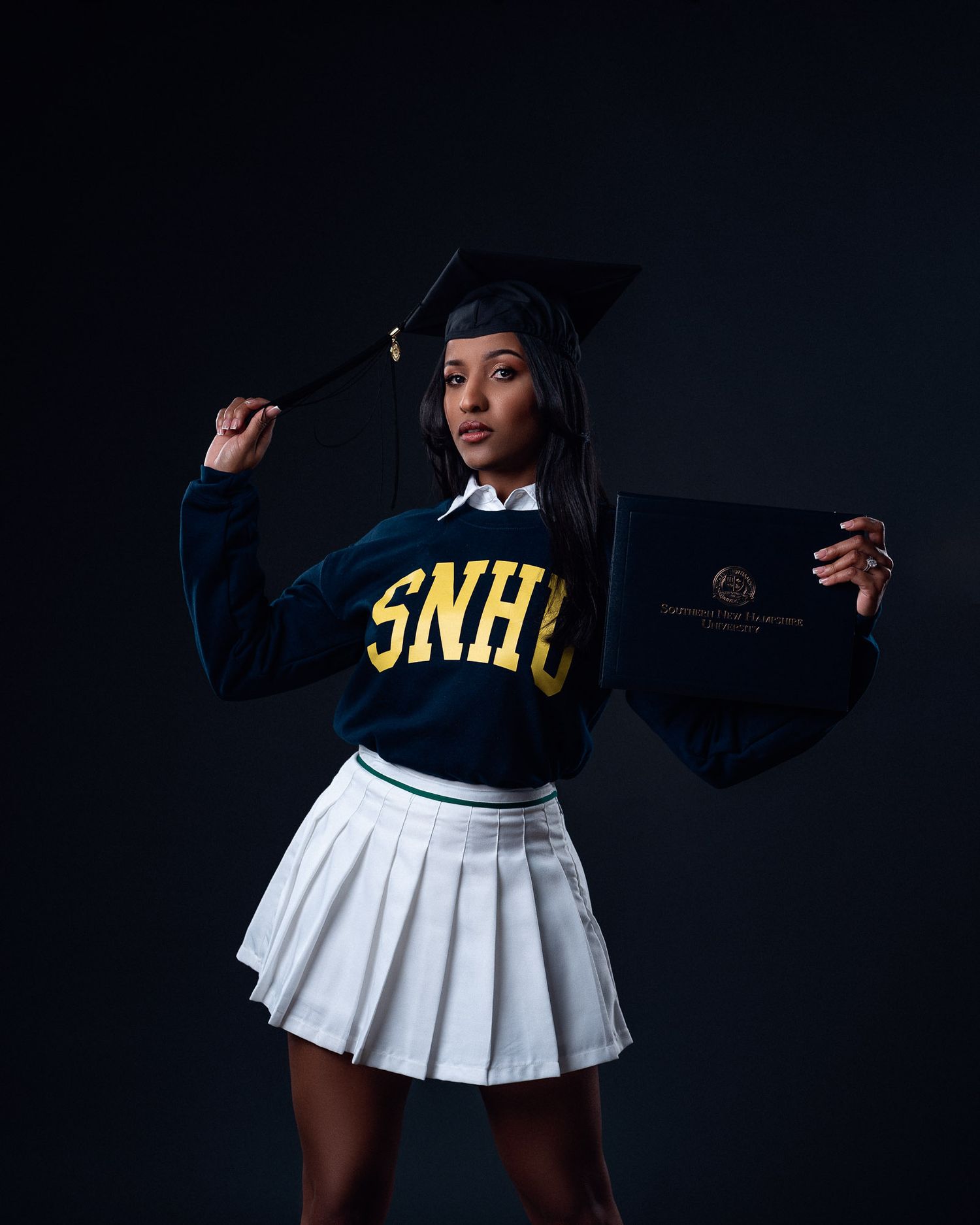 Young woman in SNHU sweater, graduation cap, and white pleated skirt holds diploma against dark background.