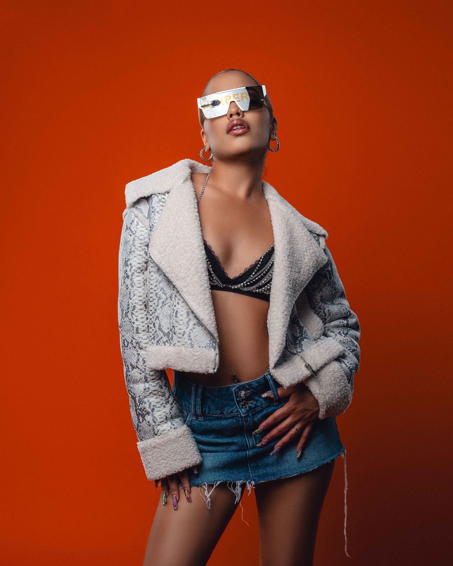 Woman in oversized shearling jacket and denim mini skirt poses against vibrant orange background.