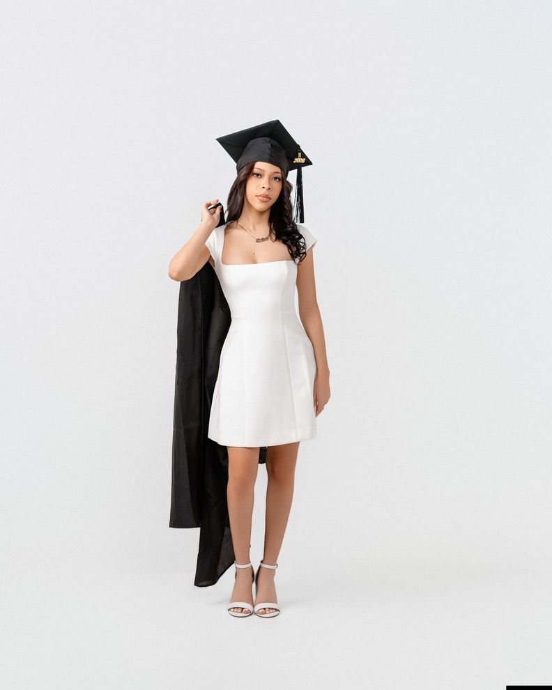 Young woman in white mini dress and black graduation cap holds gown, posing for grad photos on white background.