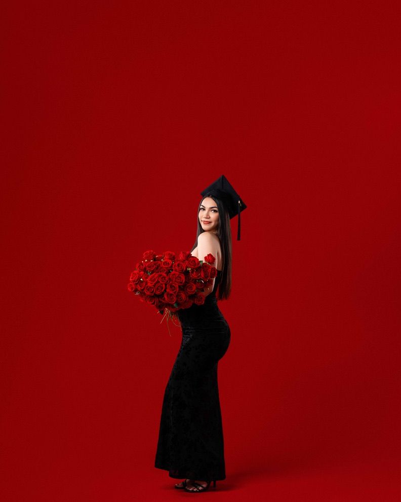 Young woman in black gown and graduation cap holding red roses bouquet against vibrant red background.