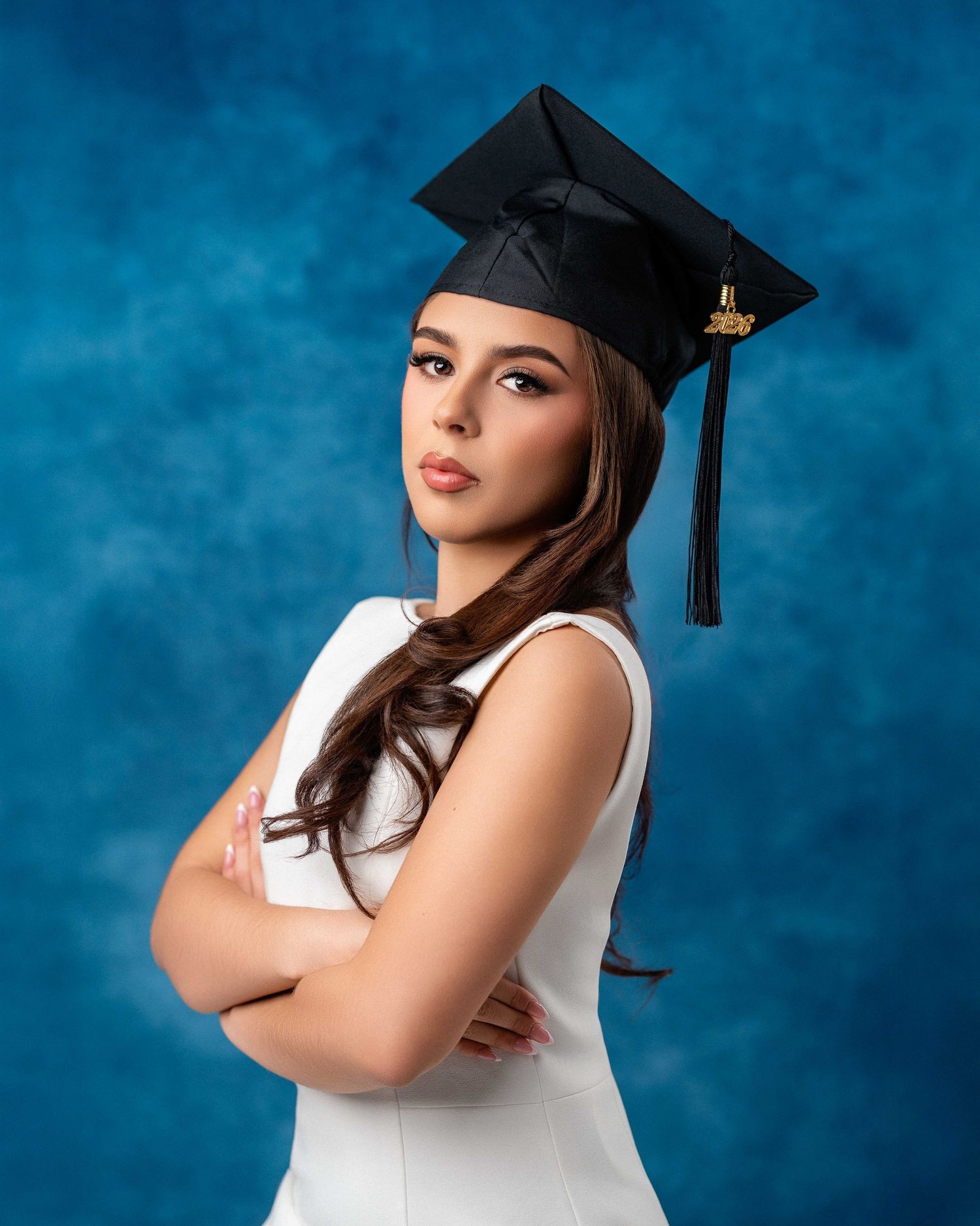 Young woman in white dress and black graduation cap poses confidently with arms crossed against blue backdrop.