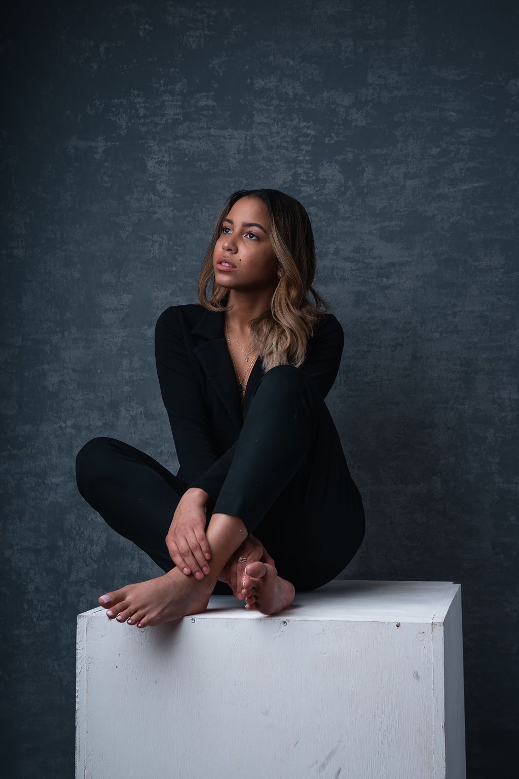 Woman in black outfit sitting cross-legged on a white cube against a dark textured background.